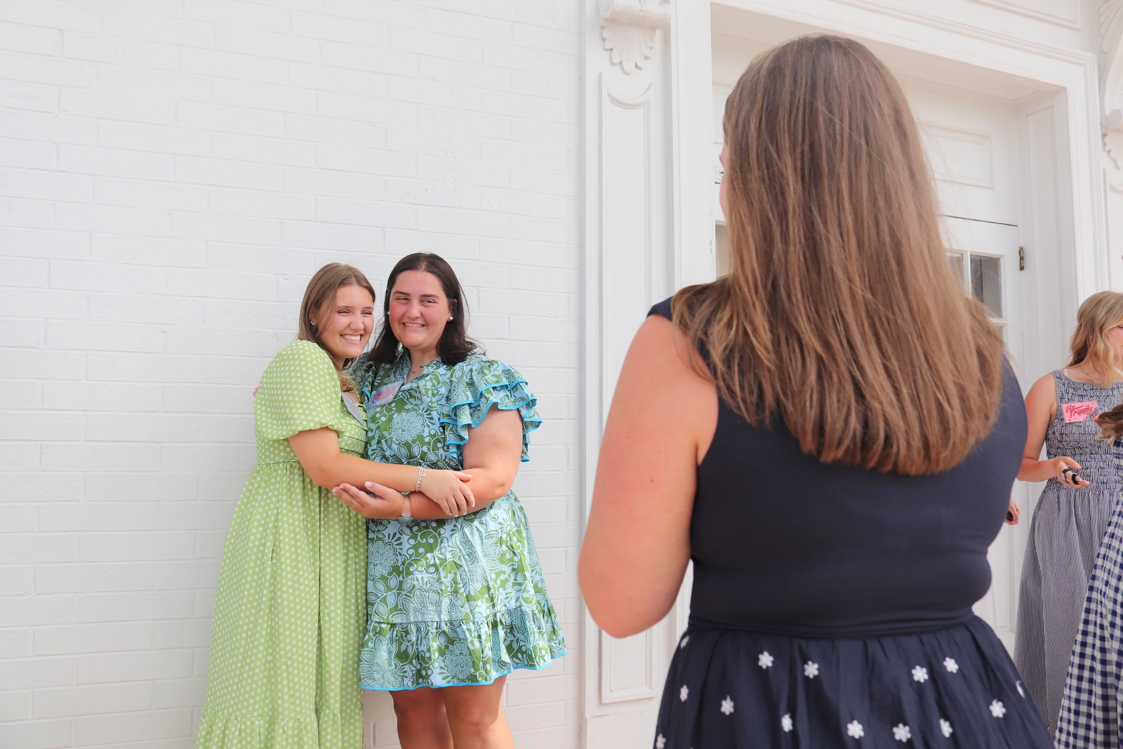 Two smiling young women in colorful dresses embrace for a photo against a white brick wall, with another woman viewing them from the foreground.