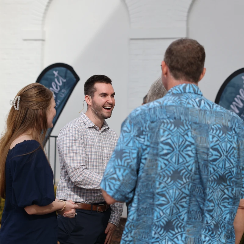 A smiling man in a plaid shirt talks with a man and a woman outdoors. Other people are in the background, mingling near a white building.