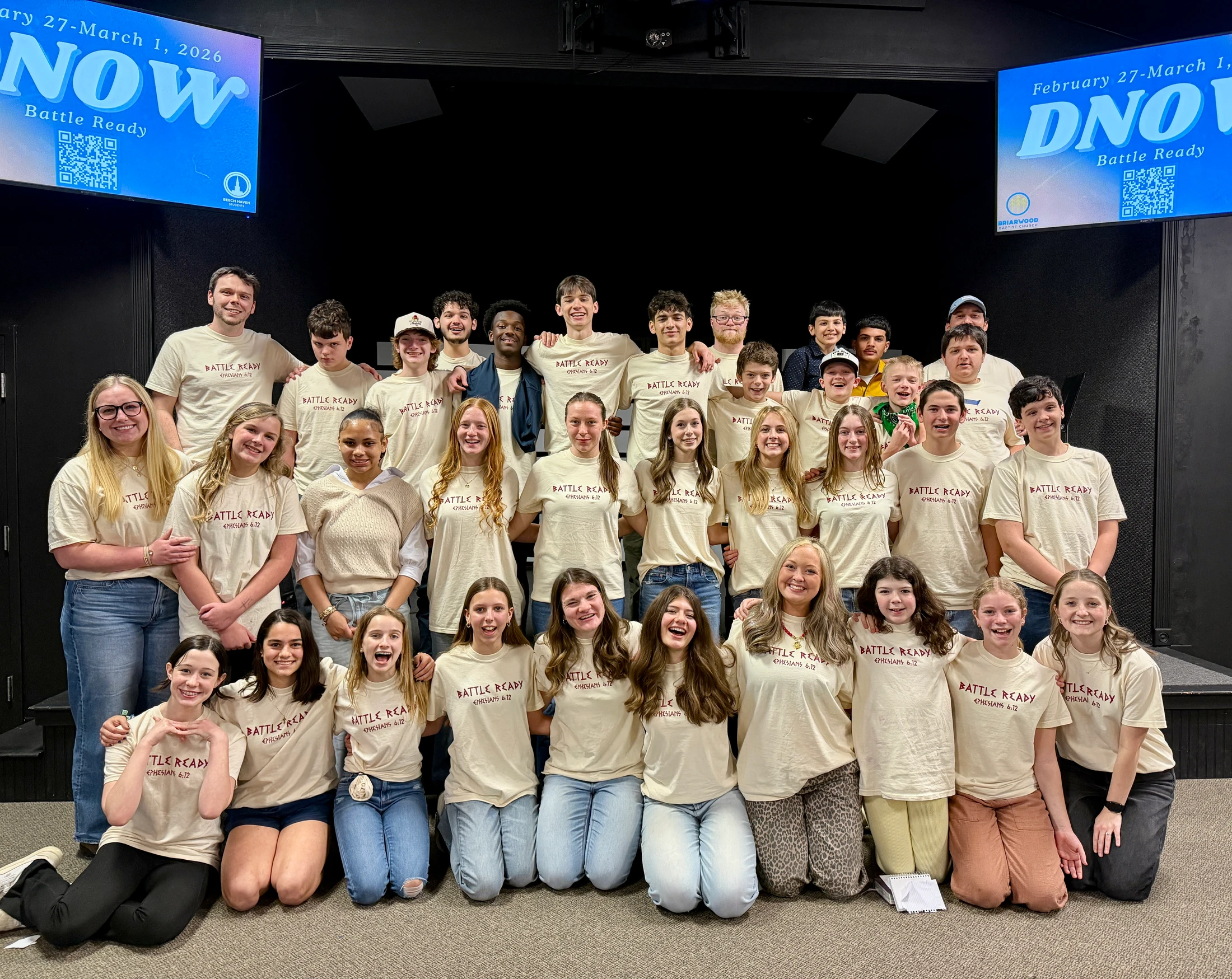 A large, diverse group of smiling teenagers and young adults pose for a photo in matching cream-colored 'Battle Ready' t-shirts in front of a dark stage.