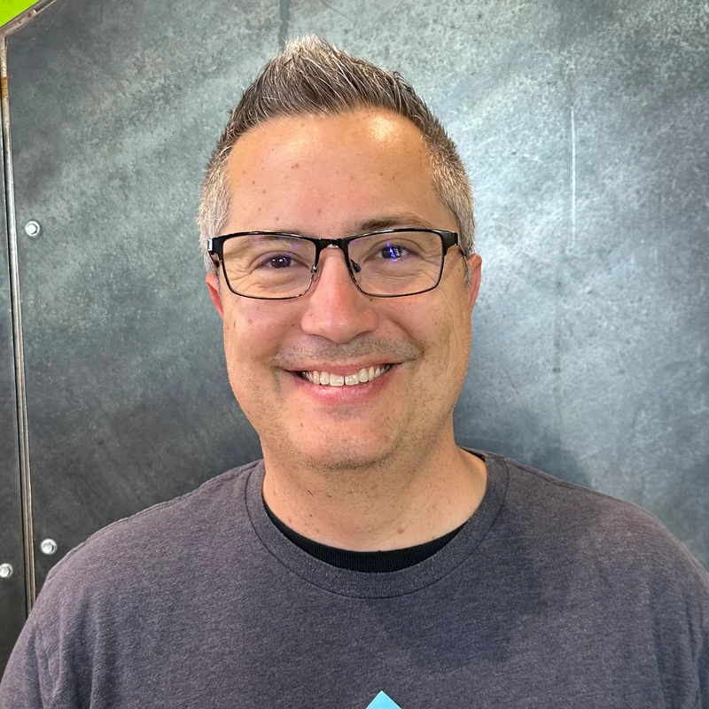 A headshot of a smiling man with short salt-and-pepper hair and black-rimmed glasses, wearing a grey t-shirt against a grey industrial background.