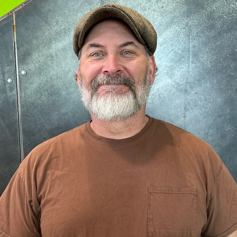 A headshot of a middle-aged man with blue eyes and a gray beard. He is smiling and wearing a brown tweed flat cap and a brown t-shirt.