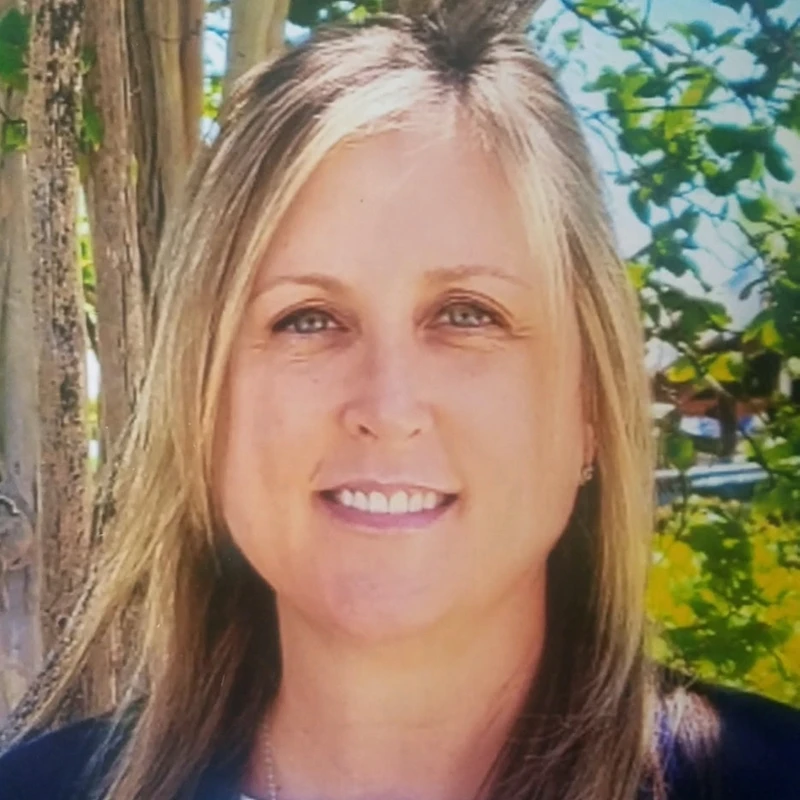 A close-up headshot of a smiling woman with shoulder-length blonde hair and light eyes. She is standing outside with trees and greenery in the background.