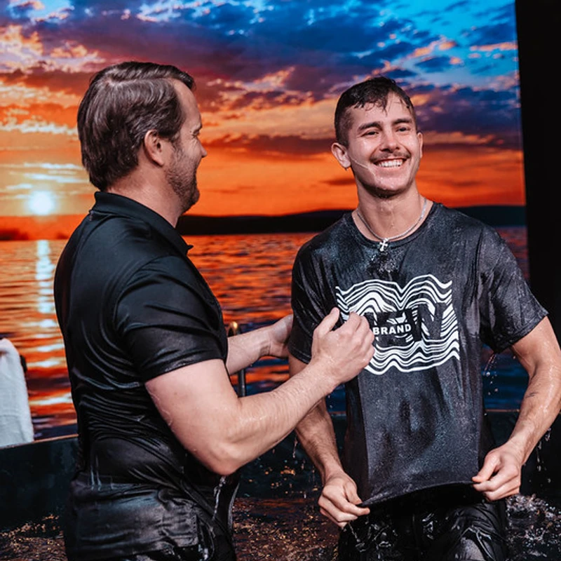 A smiling young man in a wet t-shirt stands in a baptismal font with a pastor in front of a large screen showing a beautiful sunset over water.