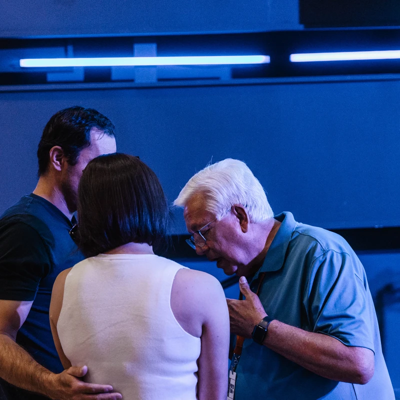 An older man with white hair prays for or counsels a man and woman who are standing with their backs to the camera in a room bathed in blue light.