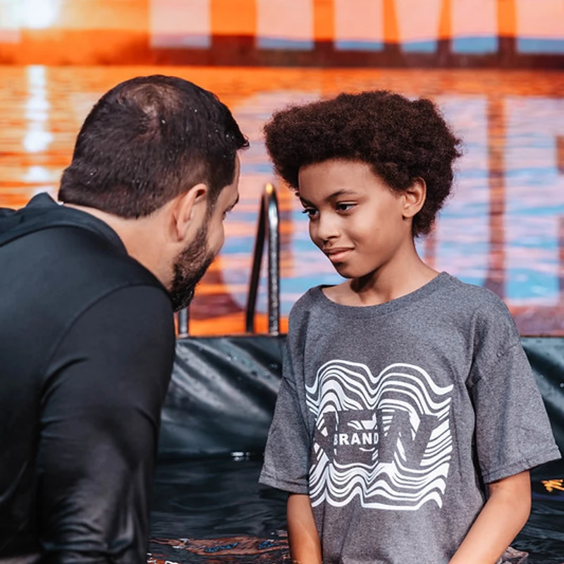 A man and a young boy stand waist-deep in a baptismal pool. The boy looks at the man with a gentle smile before the religious ceremony.