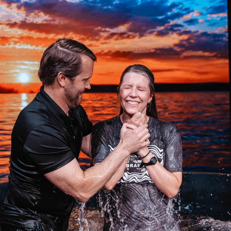A smiling pastor baptizes a woman with her eyes closed in joy. They stand in a font in front of a large screen showing a sunset over water.