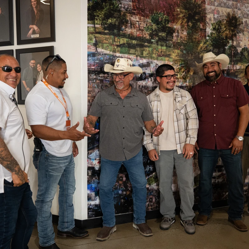 Six smiling men in casual attire, some wearing cowboy hats, stand together indoors for a group photo in front of a wall with a large photo collage.