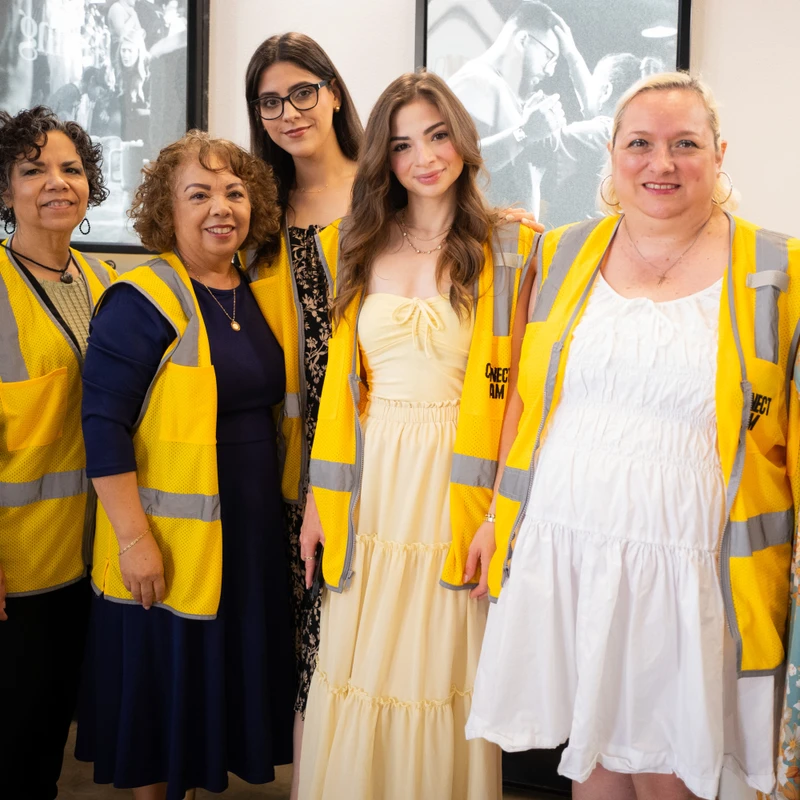 A diverse group of six women stand together and smile for a photo, with several wearing yellow and grey reflective vests over their dresses.