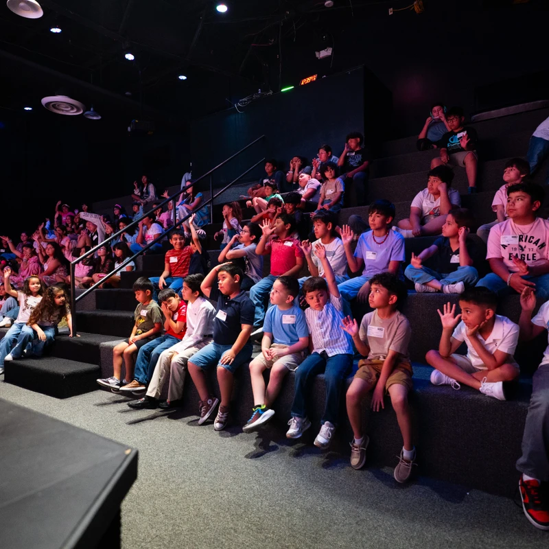 A large group of diverse children sit on tiered seating in a dark auditorium. Many have their hands raised, looking attentively towards an unseen stage.
