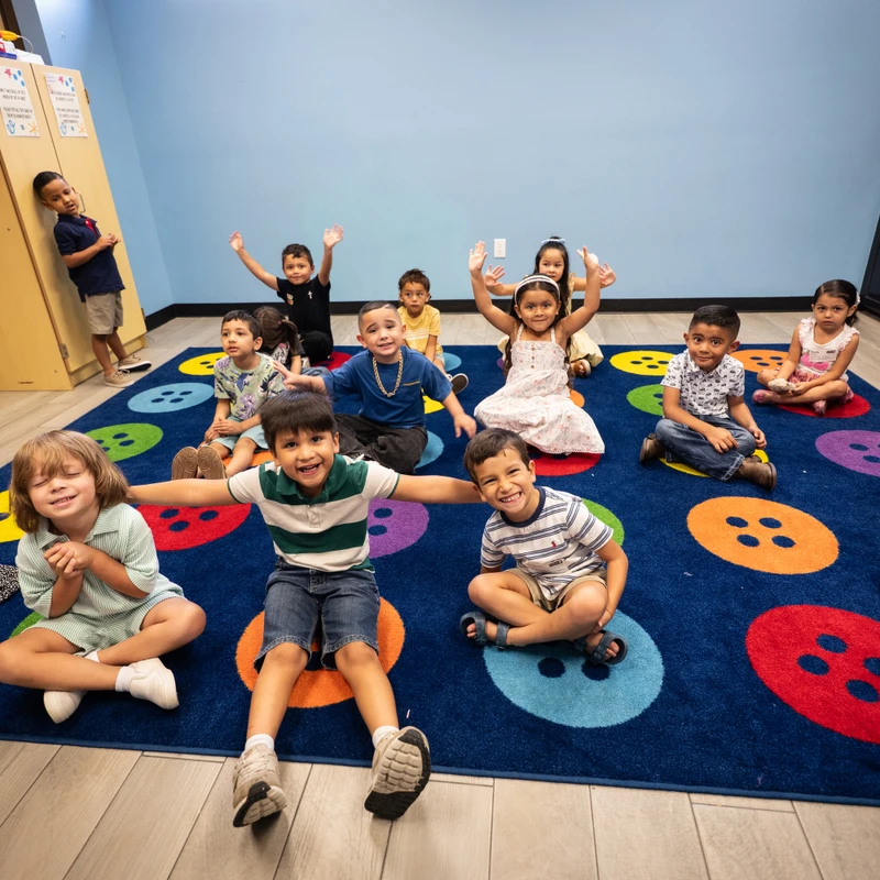 A diverse group of young children happily pose for the camera while sitting on a colorful, button-themed rug in their classroom.