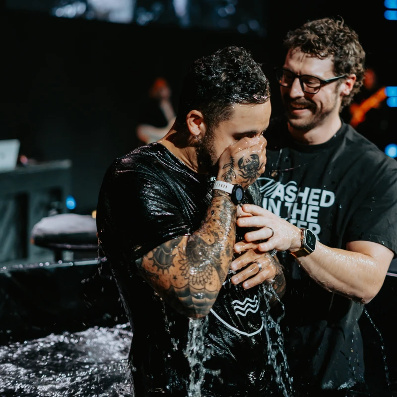 A man with tattoos on his arms wipes his face with his hand as he emerges from the water during his baptism, supported by a smiling man behind him.