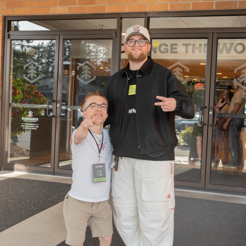 A man with dwarfism and a taller man smile and pose for a photo in front of the glass entrance to a brick building. The shorter man wears a 'Leader' name tag.