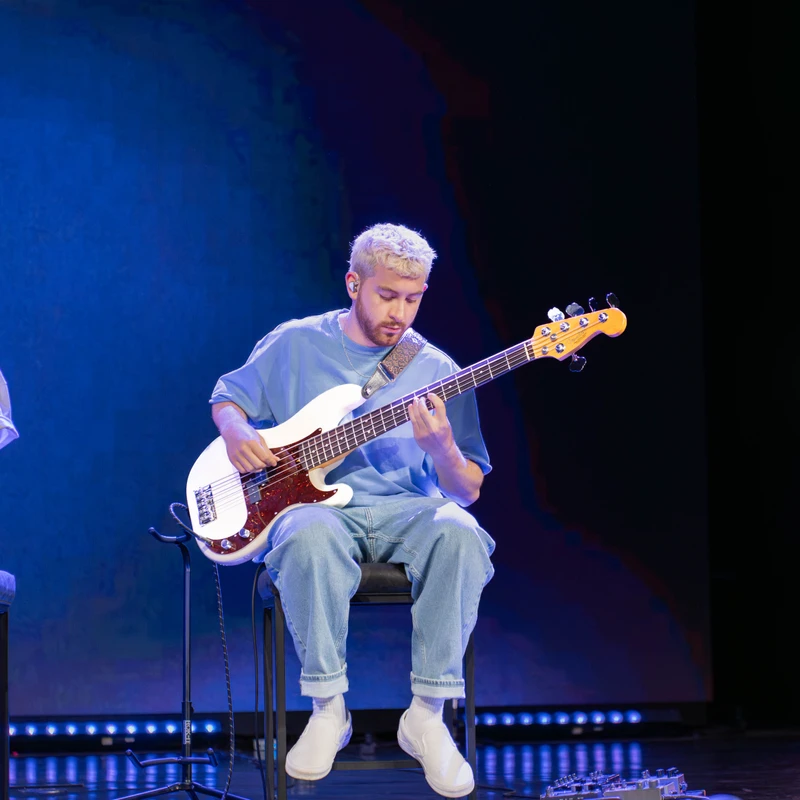 A young man with bleached hair sits on a stool, playing a white electric bass guitar on a stage with a blue background and lighting.