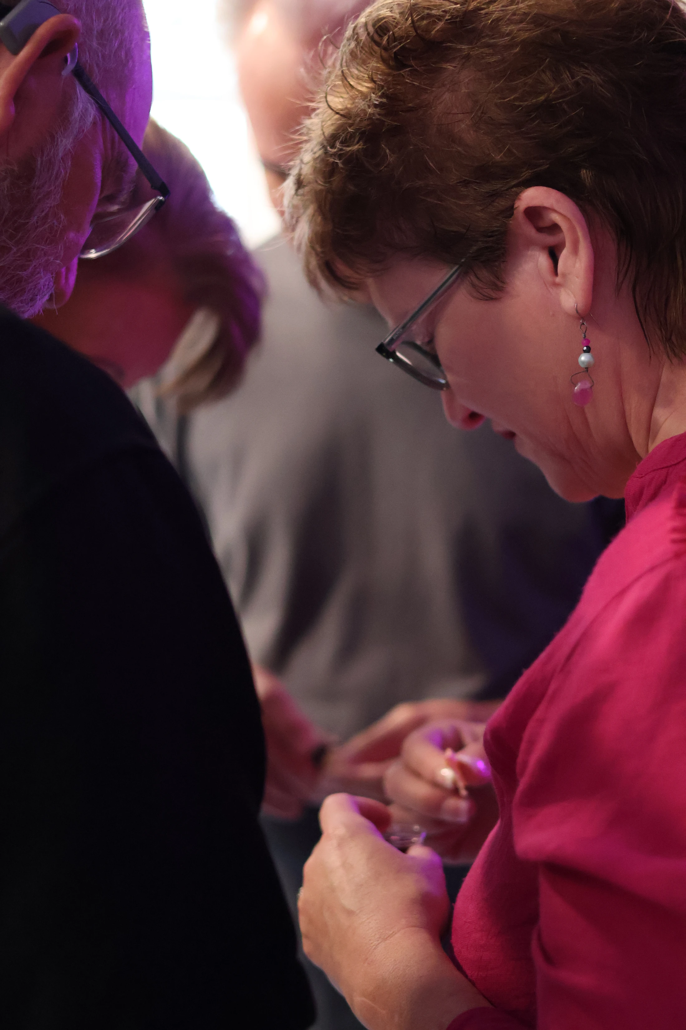 A woman in a pink shirt and glasses looks down in concentration, examining something in her hands. Two other people are partially visible around her.