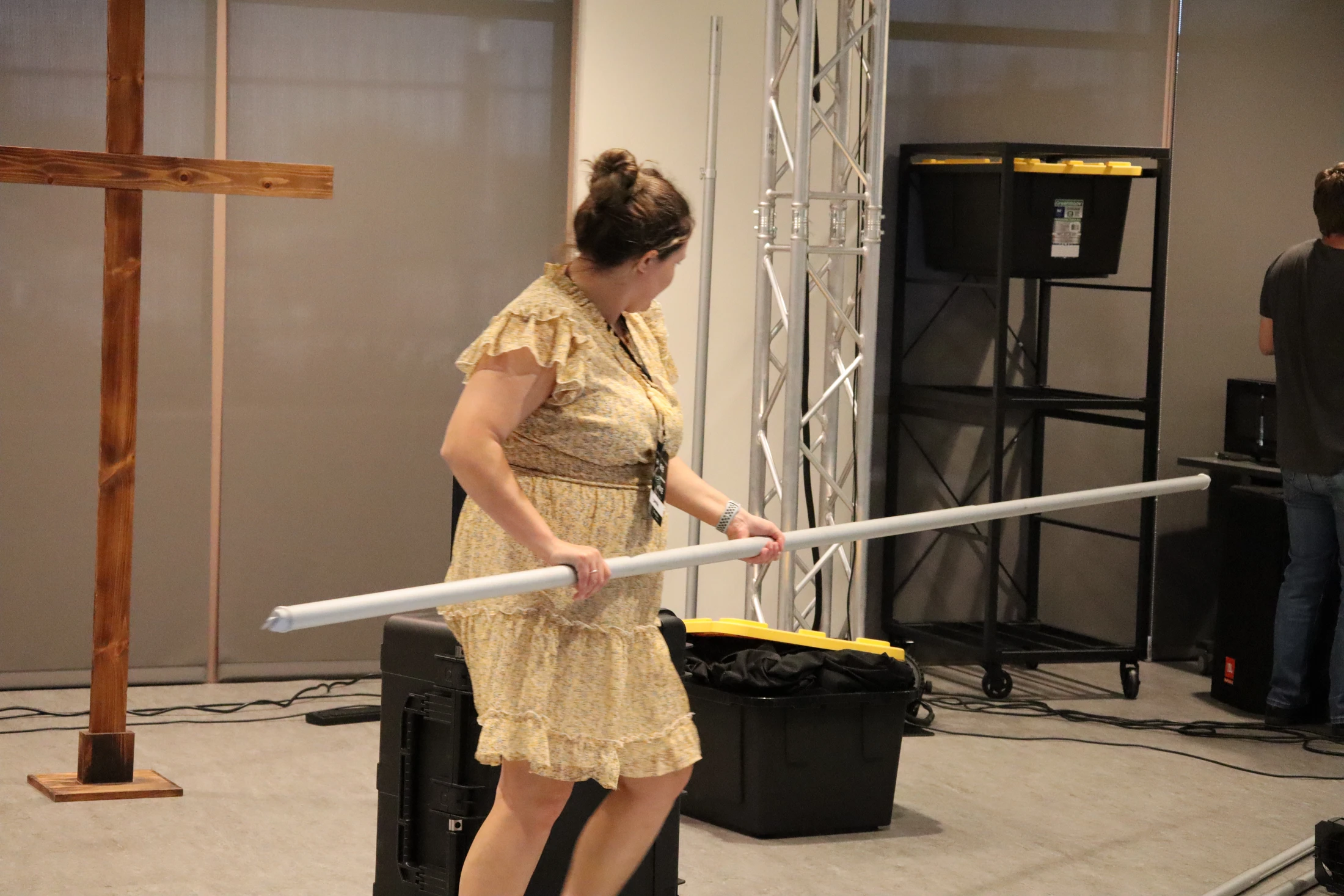 A woman in a yellow floral dress carries a long white pole, helping set up backstage where a wooden cross and stage equipment are visible.