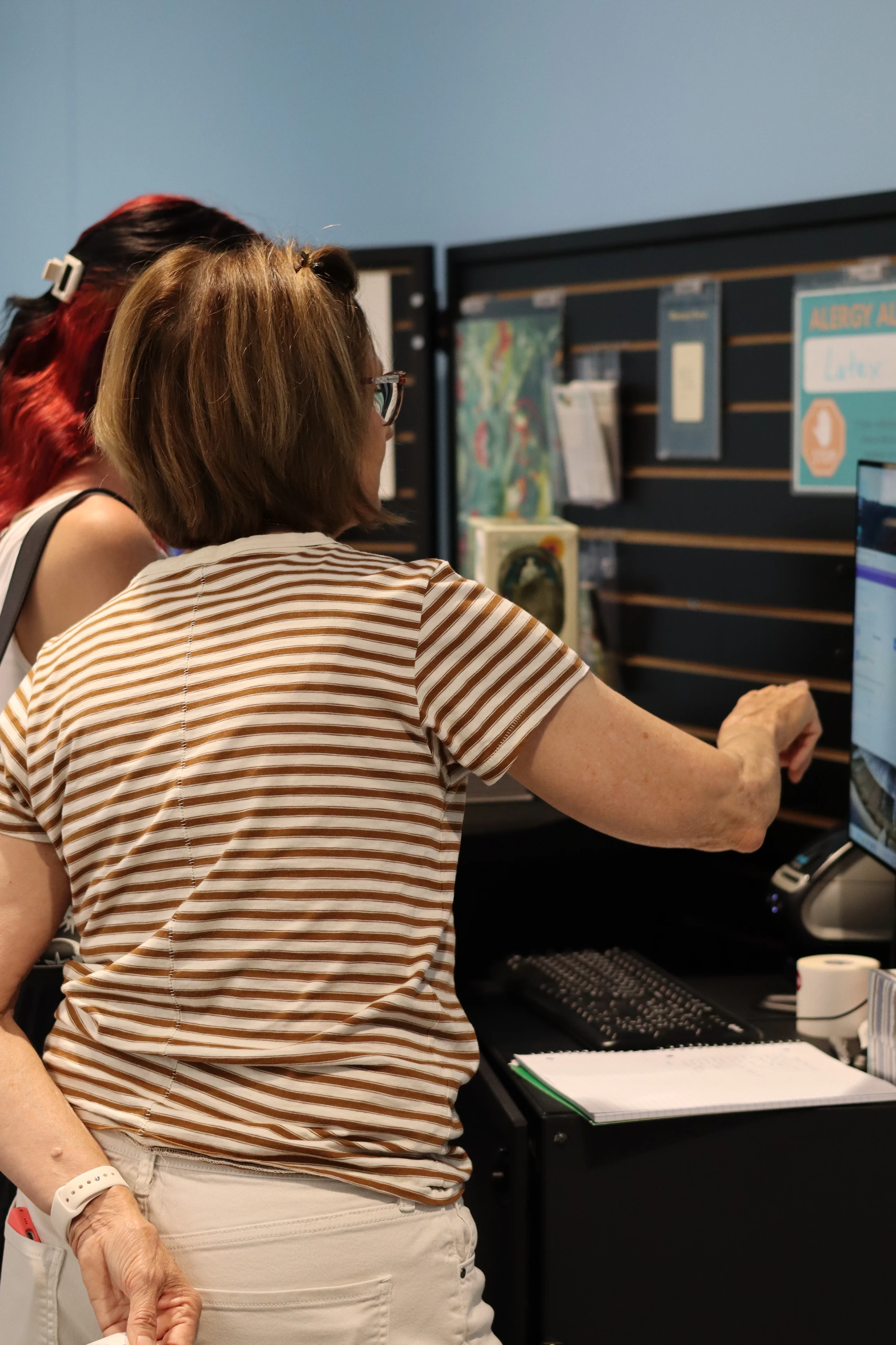 A woman in a striped shirt, seen from behind, points to a computer screen on a desk while standing next to another person.