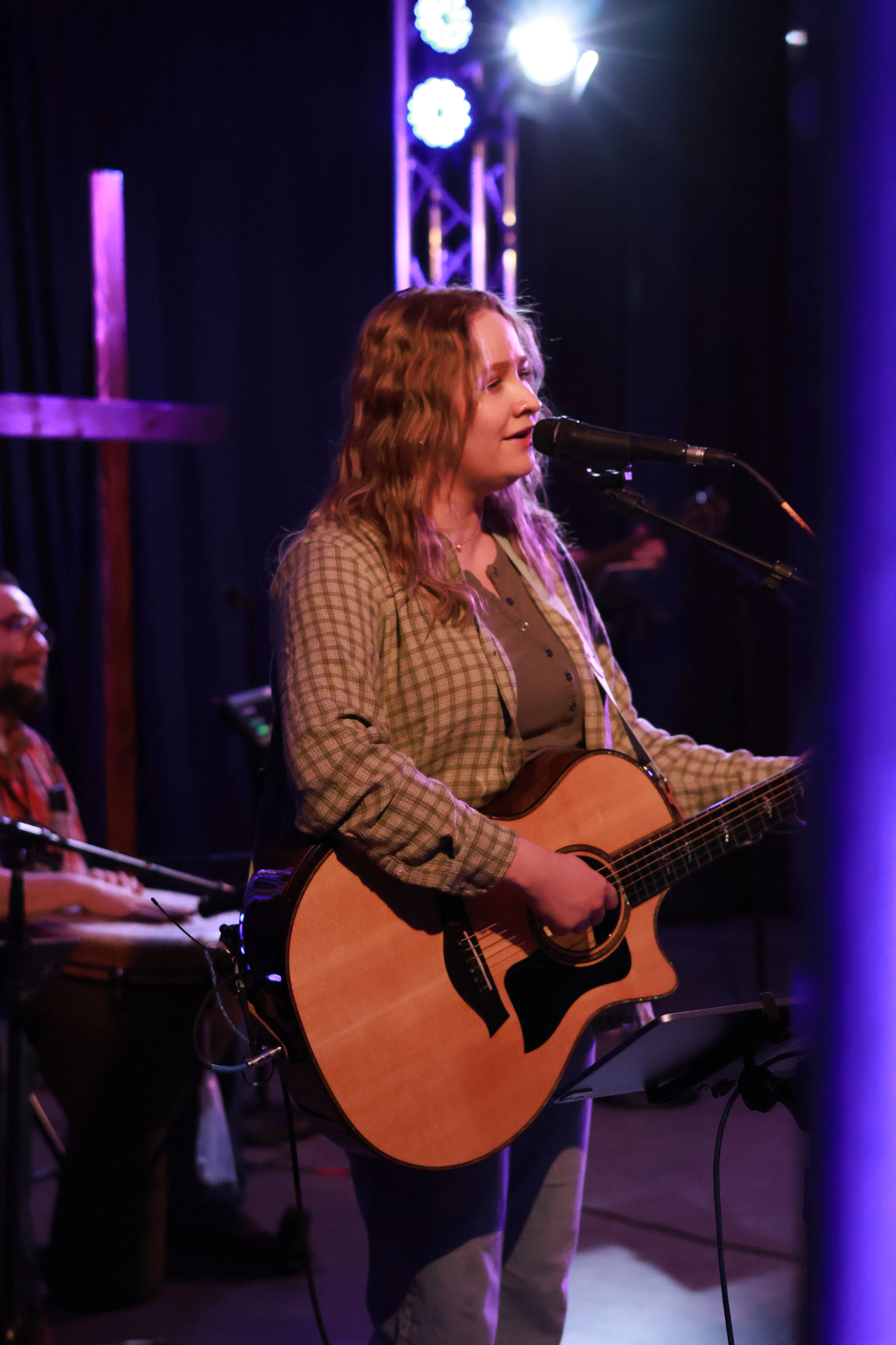 A woman with wavy blonde hair sings into a microphone while playing an acoustic guitar on a dimly lit stage with purple lights and a cross in the background.