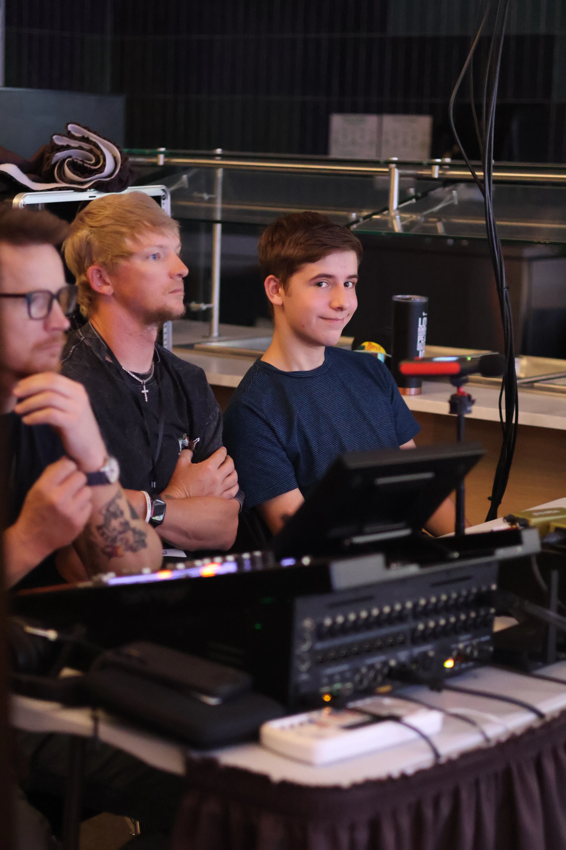 A young man in a blue t-shirt smiles at the camera while sitting between two other men behind a large audio mixing console at an event.