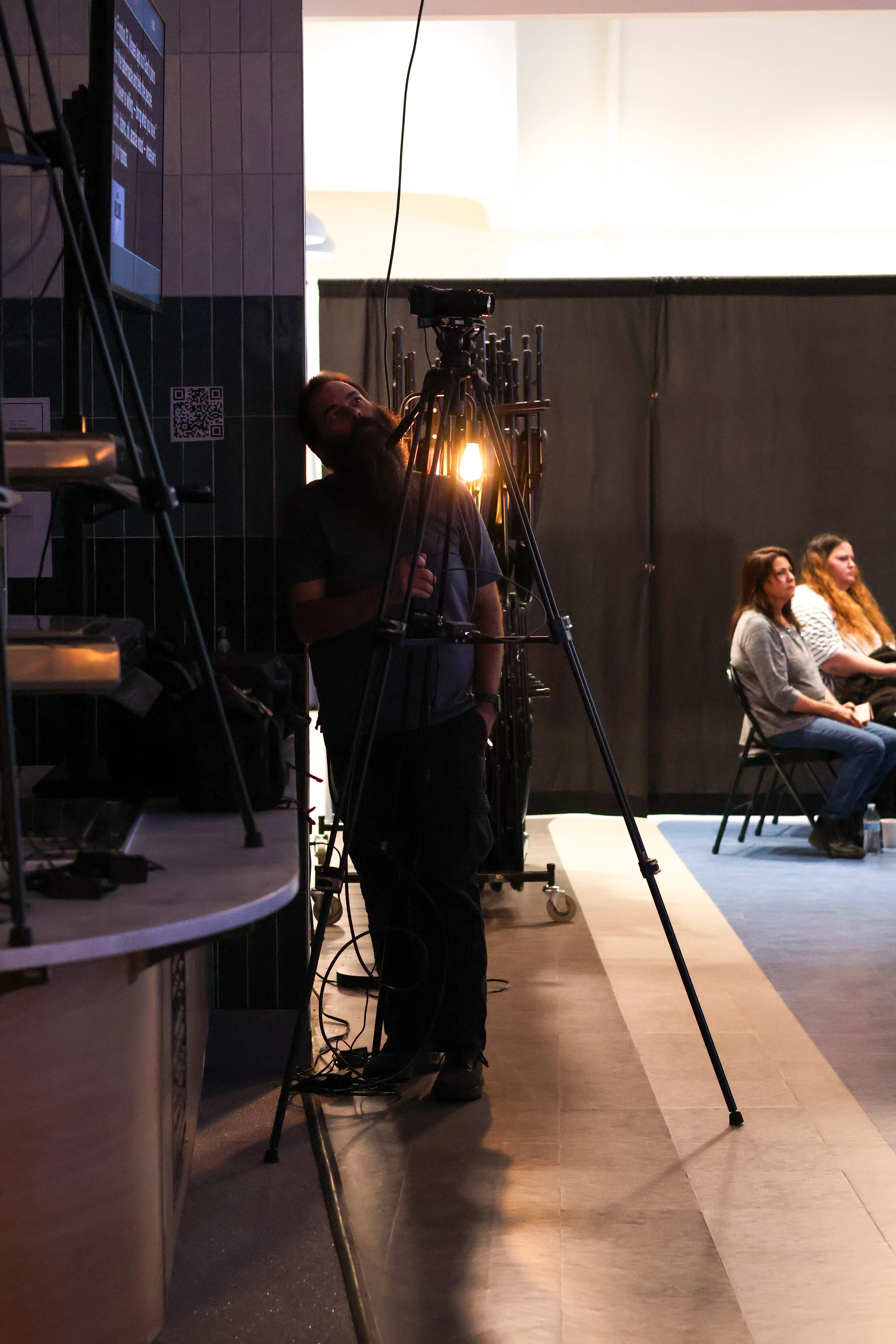 A bearded man stands behind a camera on a tripod, illuminated by a warm light. In the background, two women sit in an audience watching.