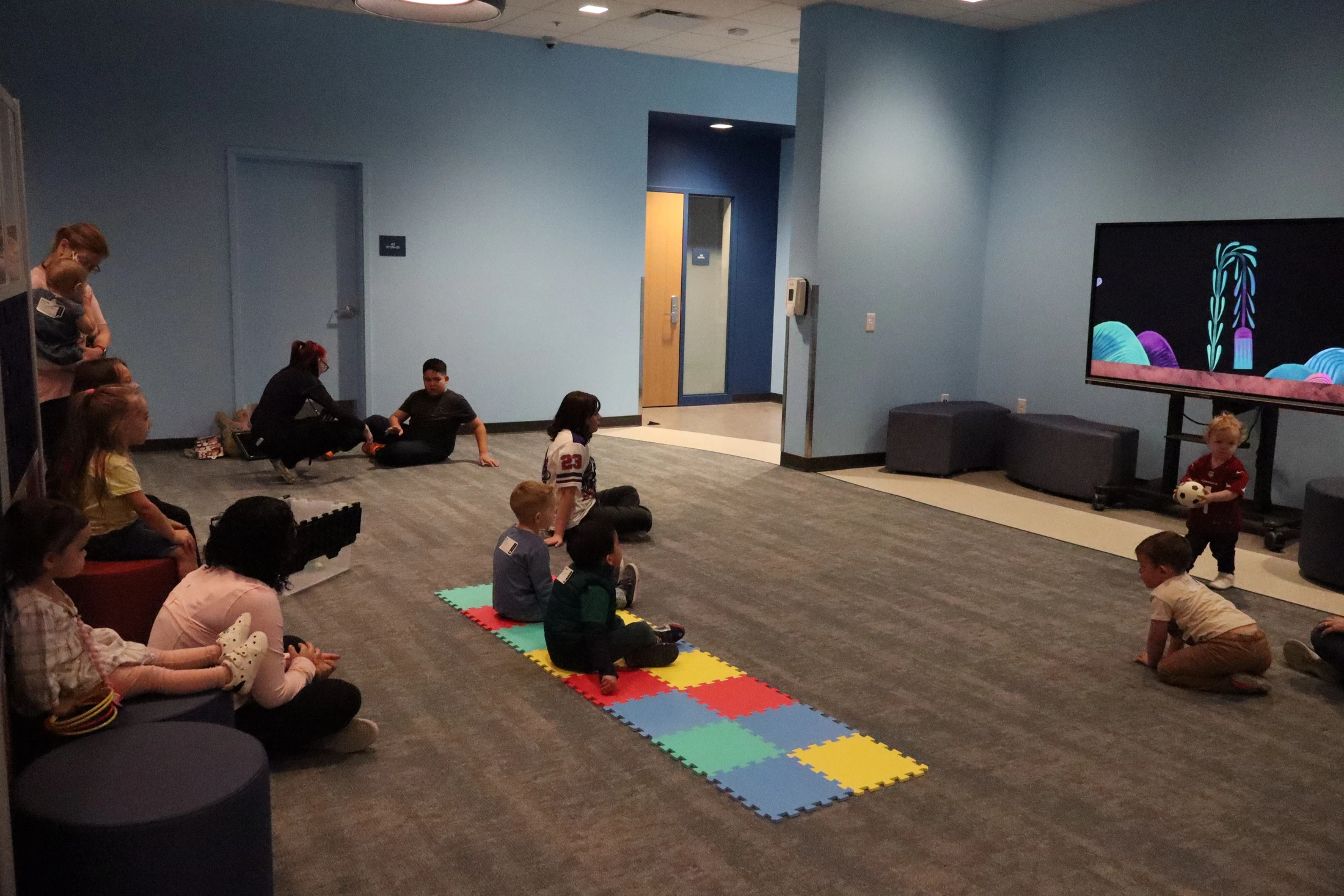 A group of young children and several adults sit on the carpeted floor of a large room, watching a colorful cartoon on a large screen.
