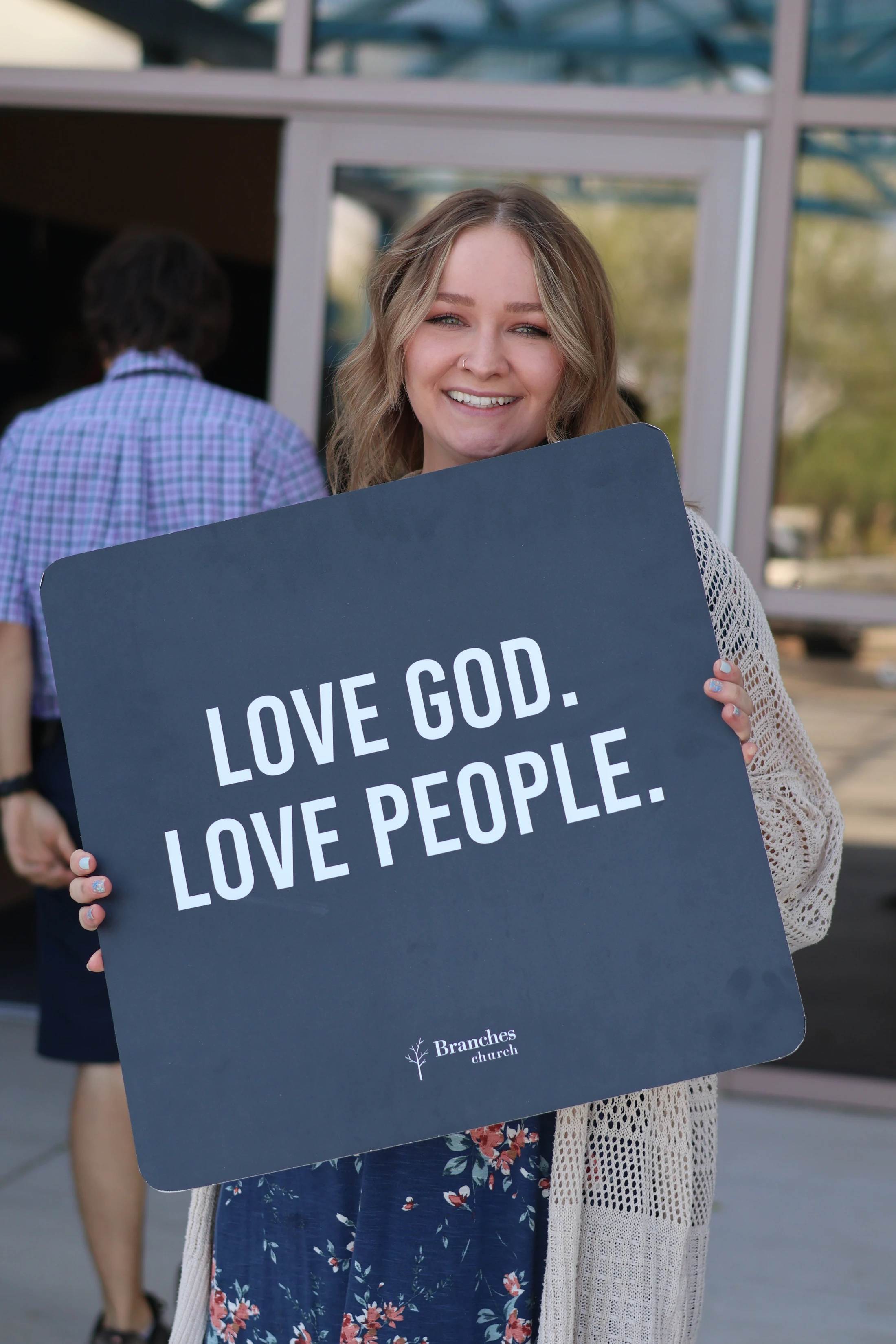 A smiling young woman with blonde hair holds a sign that reads 'LOVE GOD. LOVE PEOPLE.' outside the entrance of a building.