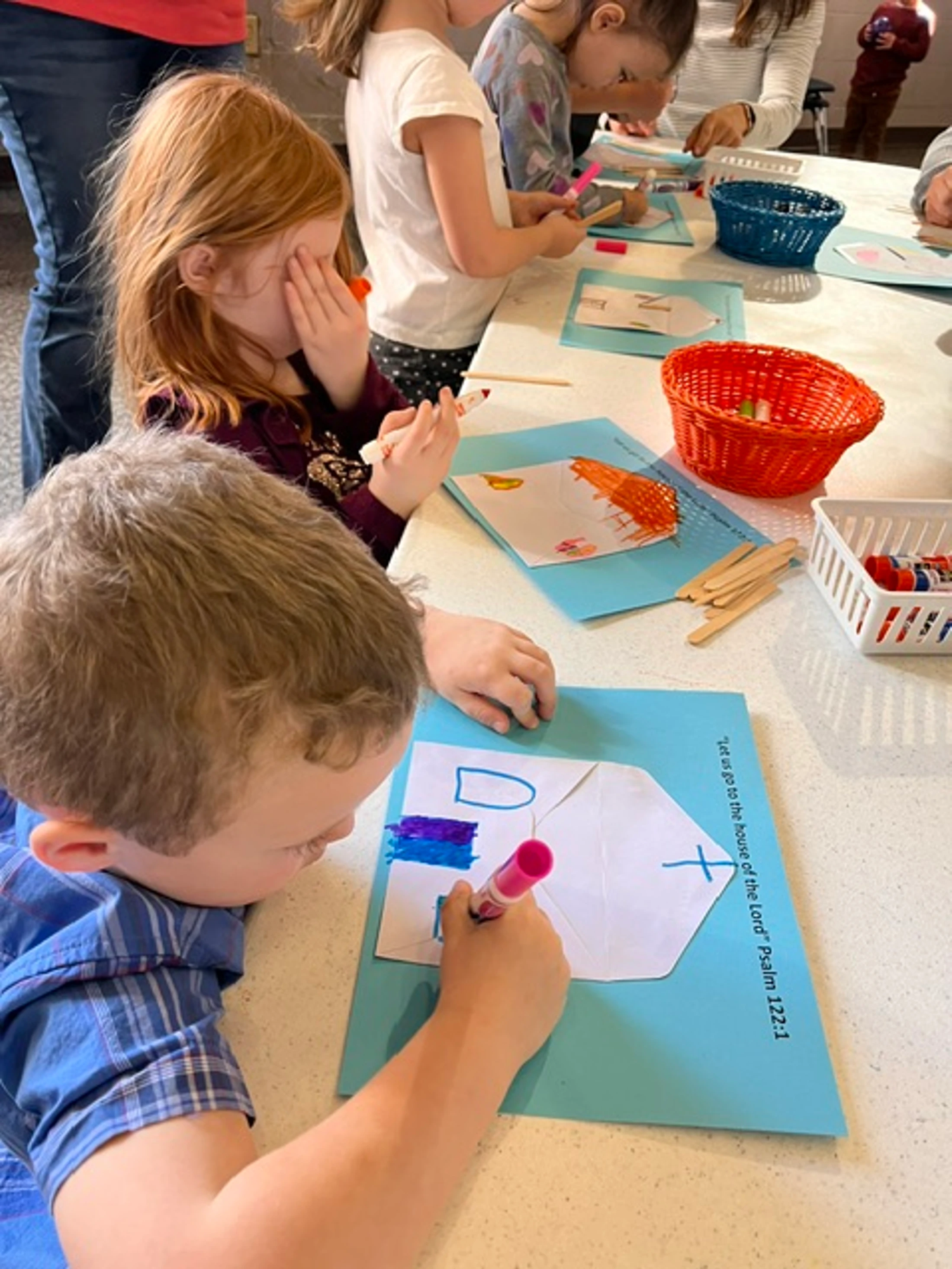 A group of young children sit at a long table coloring and creating crafts with paper, markers, and popsicle sticks in a classroom setting.