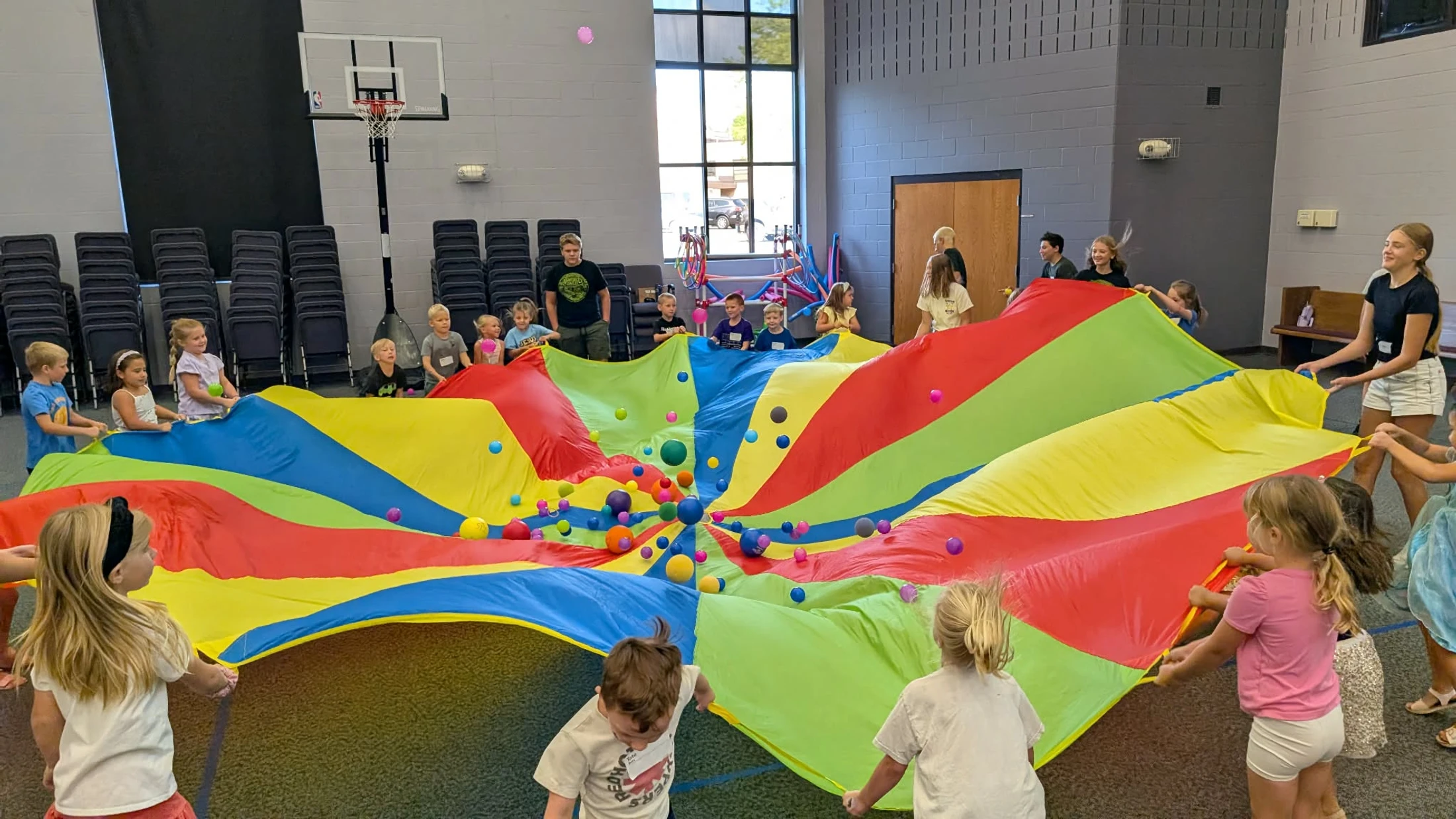 A group of children in a large room stand in a circle, holding the edges of a large, colorful parachute with bouncing balls on it.