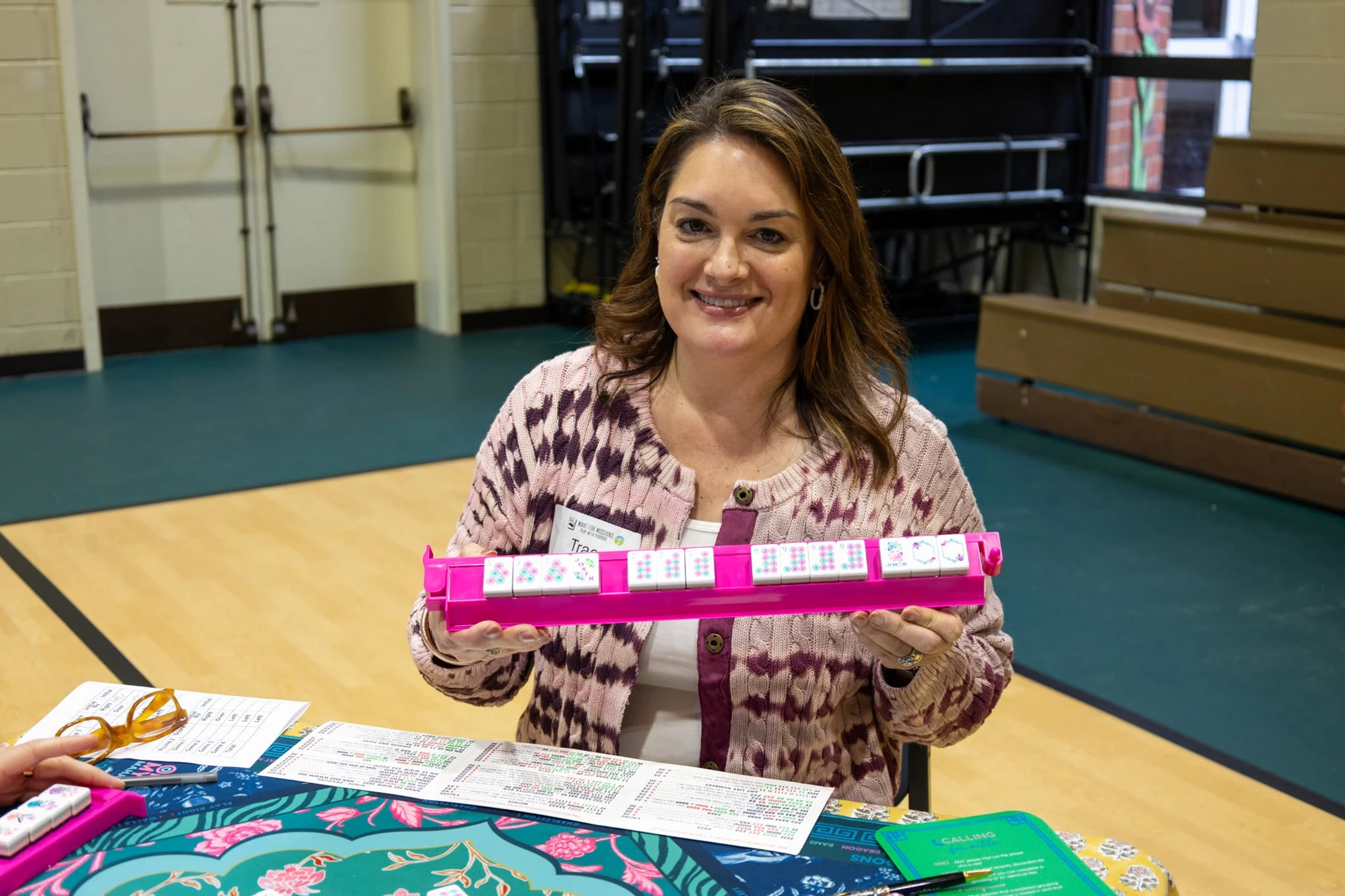 A woman in a pink and purple cardigan smiles as she holds up her pink rack of mahjong tiles while sitting at a game table.