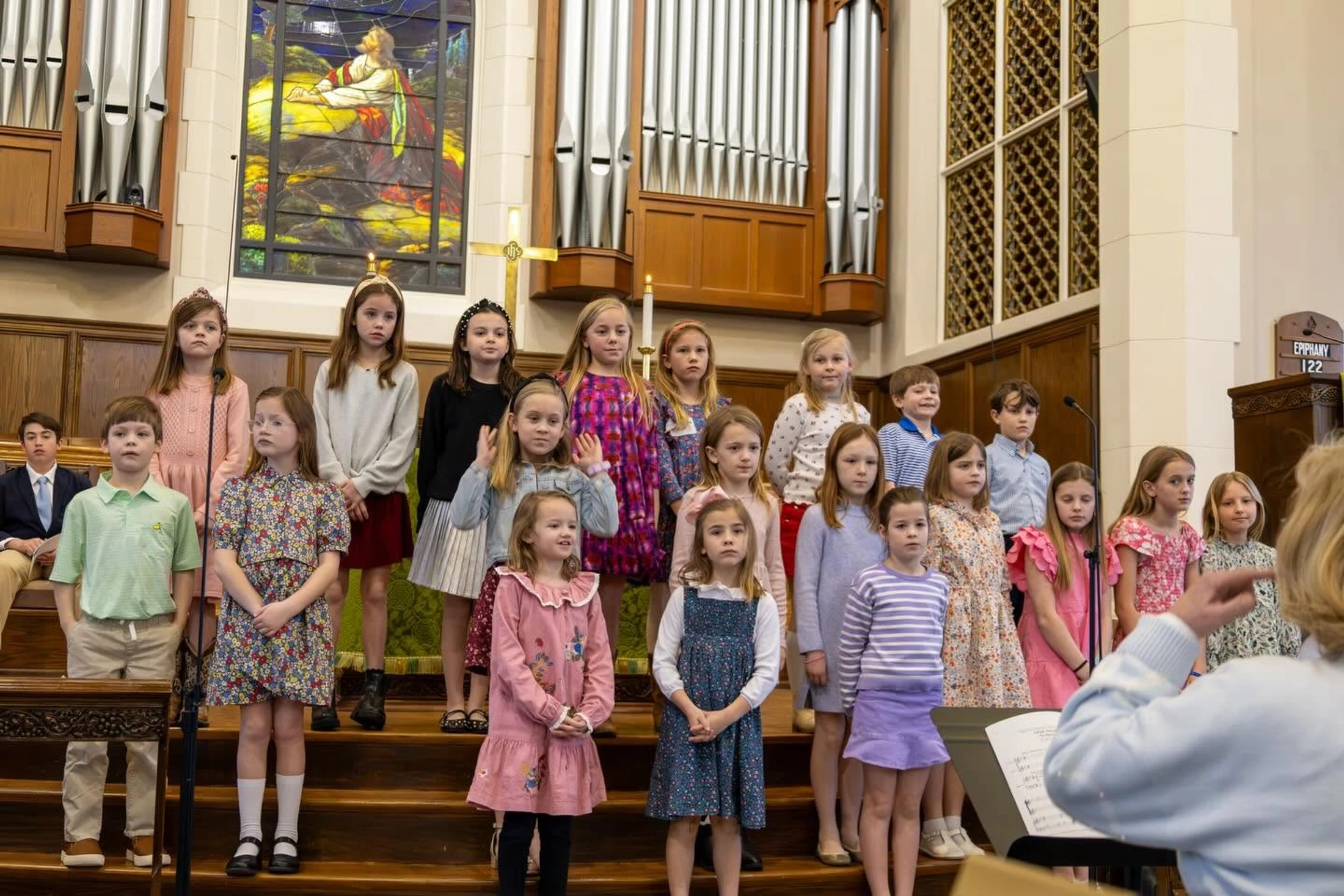 A group of young children in a choir stand on risers in a church sanctuary, singing while a conductor leads them from the foreground.