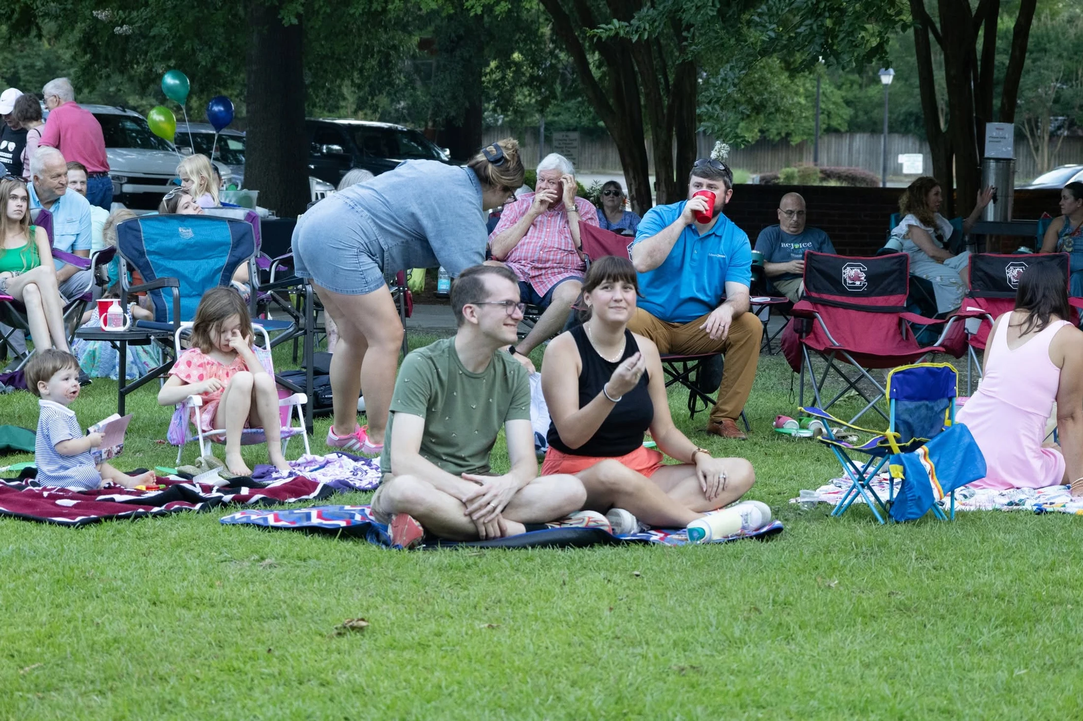A diverse group of adults and children relax on blankets and folding chairs on a green lawn, enjoying a community event outdoors under some trees.
