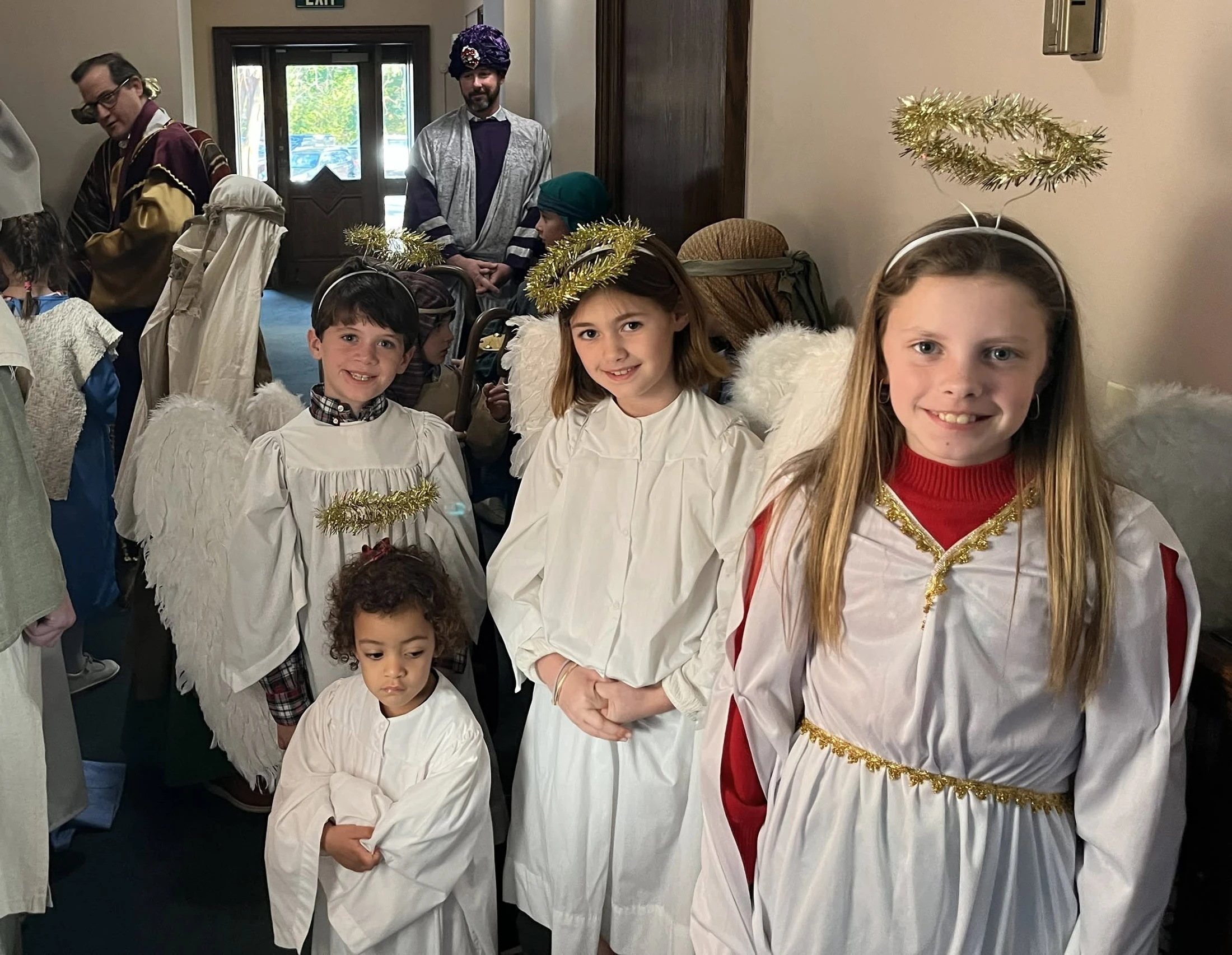 A group of children dressed in white angel costumes with wings and tinsel halos smile for a picture. Adults in nativity costumes stand in the background.