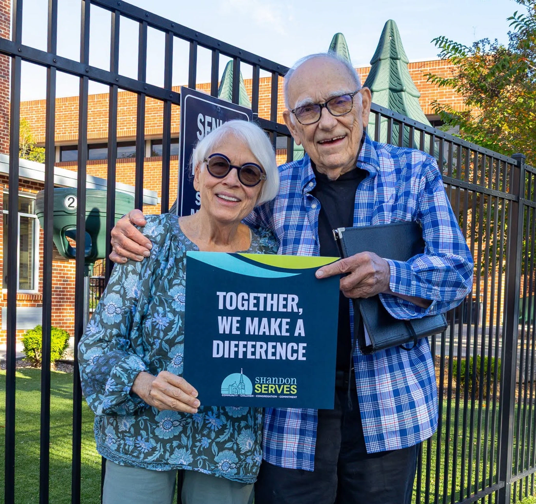 An elderly couple smiles while standing outside. The woman holds a sign that reads, "Together, We Make A Difference" for a Shandon Serves event.