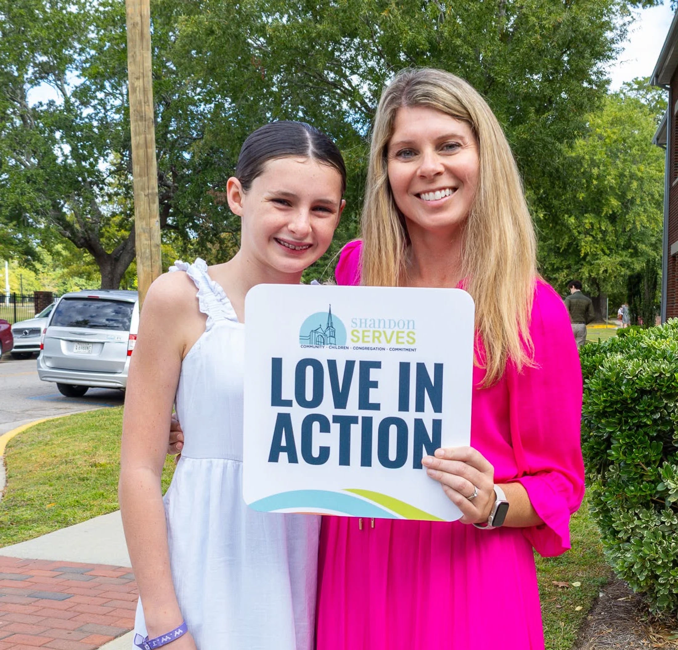 A woman in a pink dress and a young girl in a white dress smile while holding a sign that says 'Shandon Serves' and 'Love in Action'.