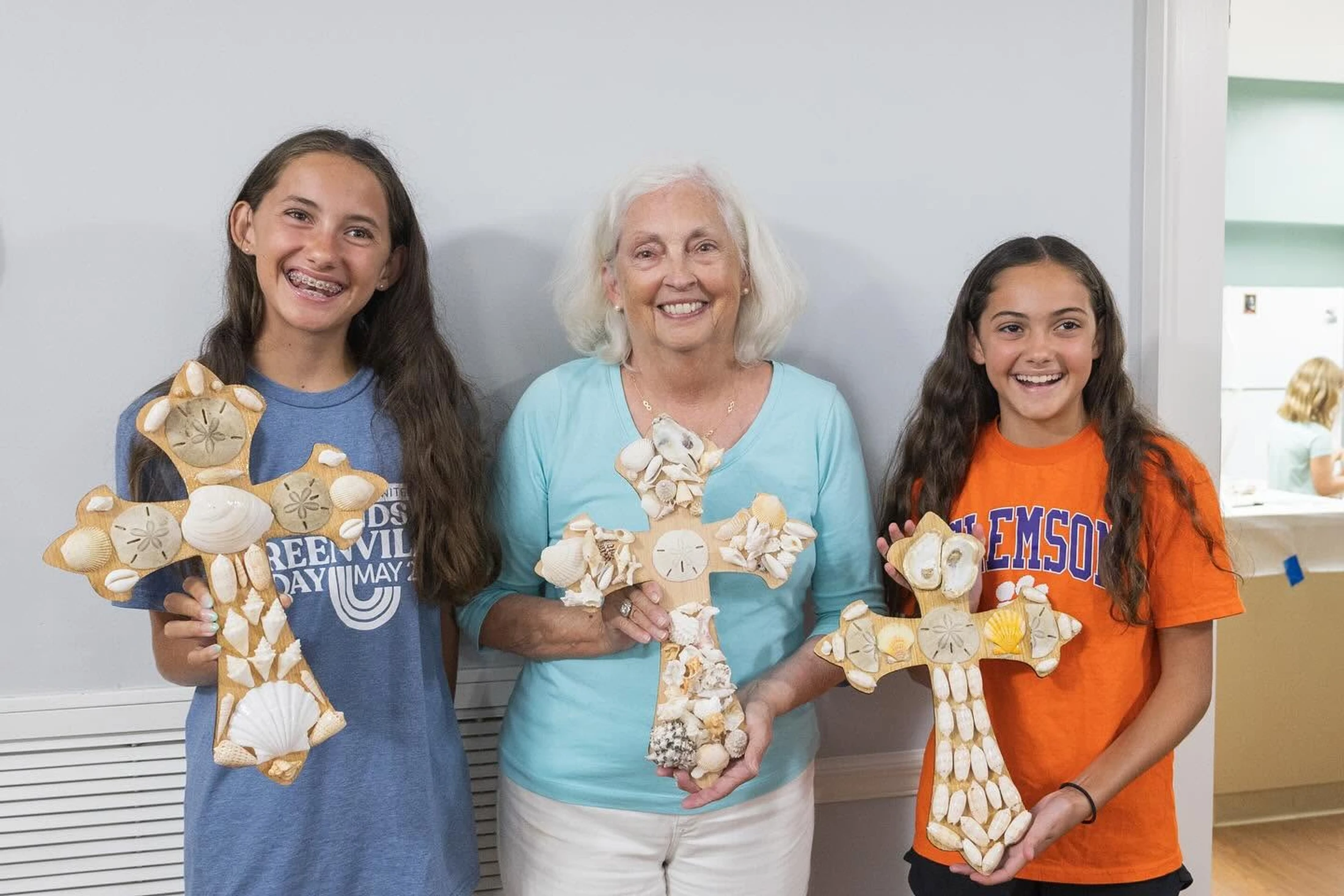 An older woman and two young girls smile as they hold up wooden crosses they have decorated with a variety of seashells and sand dollars.