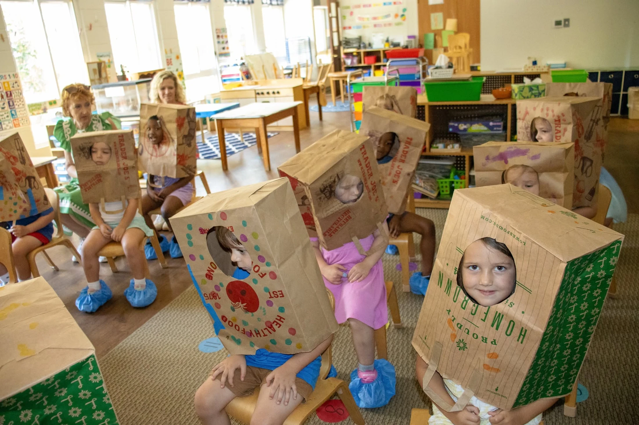A group of young children in a classroom wearing decorated paper grocery bags over their heads like helmets, with their faces peeking through cut-out holes.