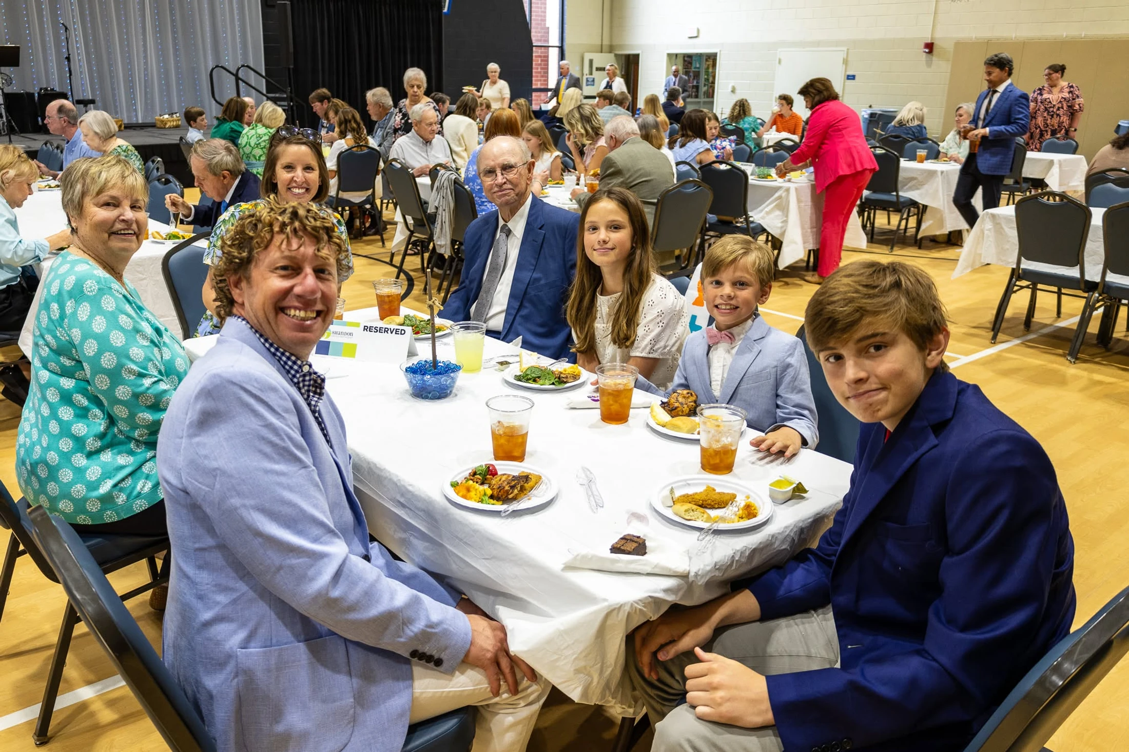 A large, multi-generational family smiles while seated at a long table with a white tablecloth, eating a meal during a banquet in a large hall.