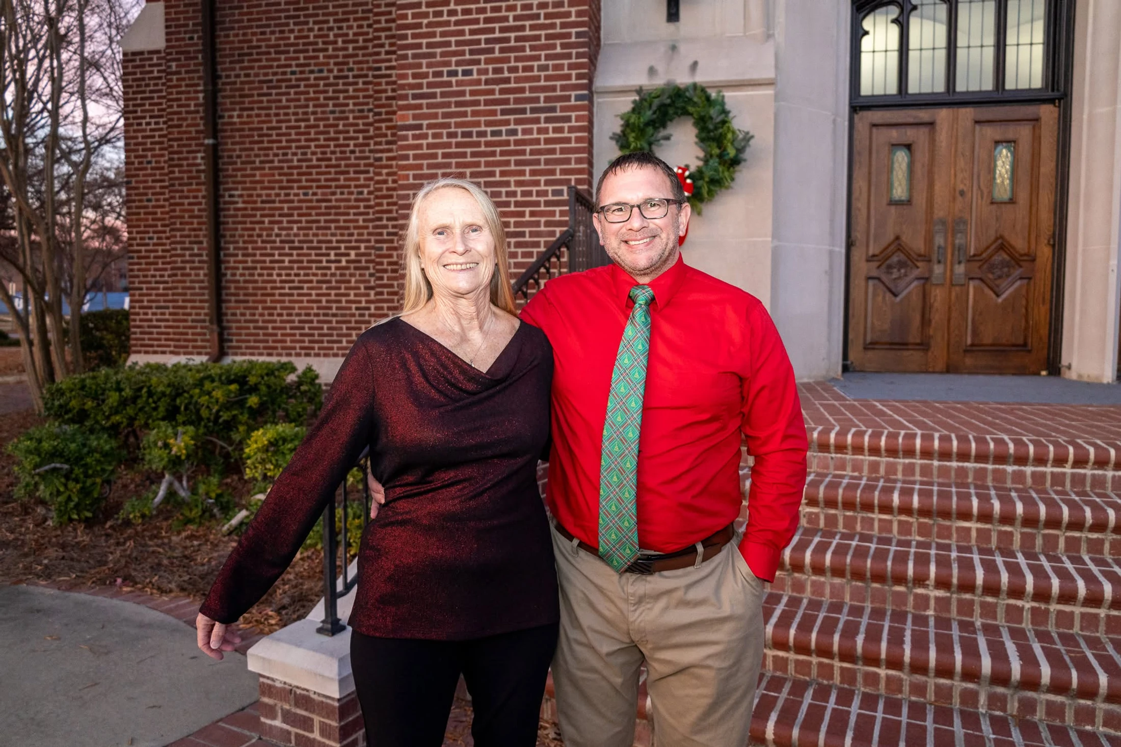 A smiling man and woman pose for a photo outside a brick church. The man is in a red shirt and festive tie, and a Christmas wreath is on the wall.
