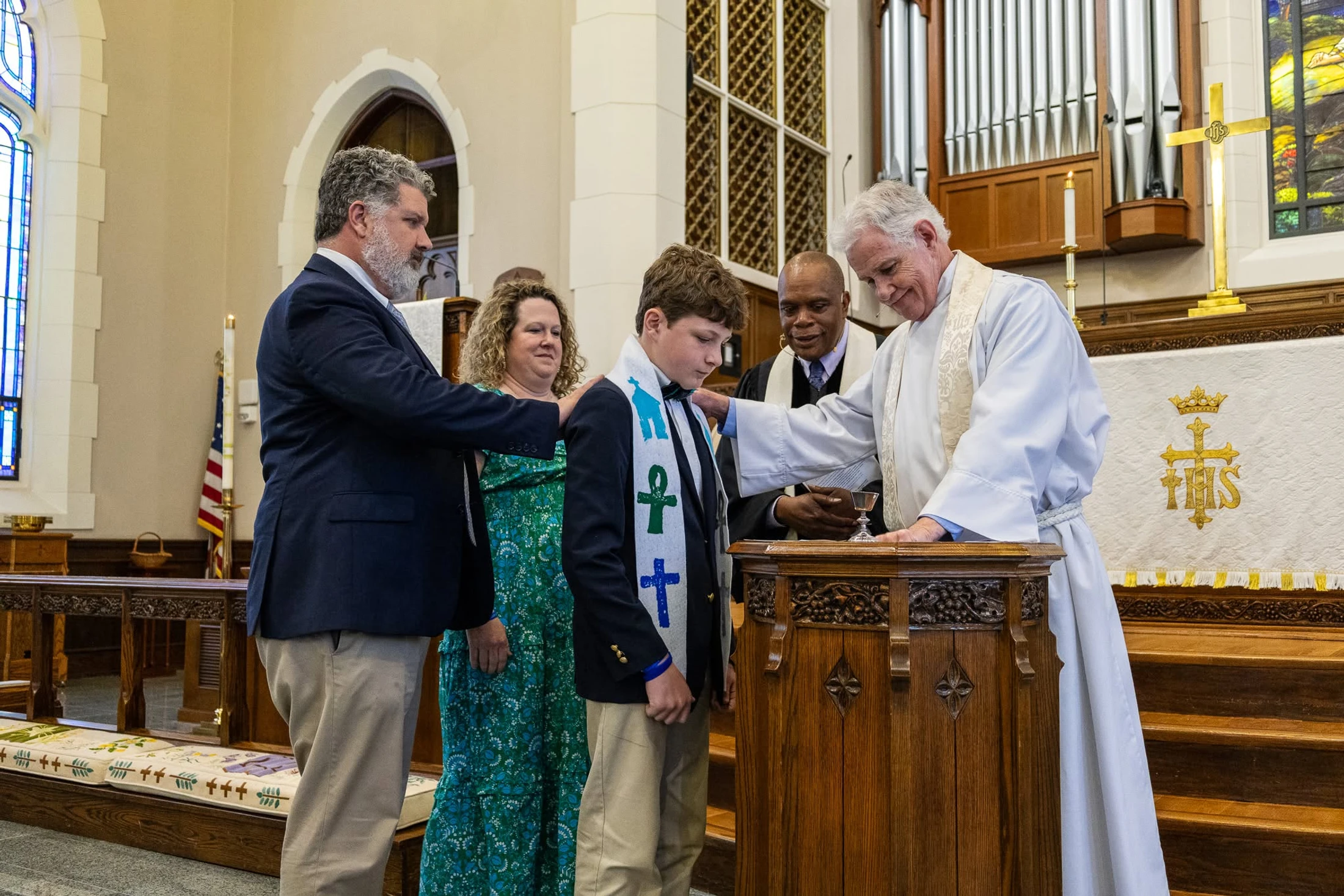 A priest blesses a young boy at an altar during a church ceremony, with his parents and another clergyman standing beside them.