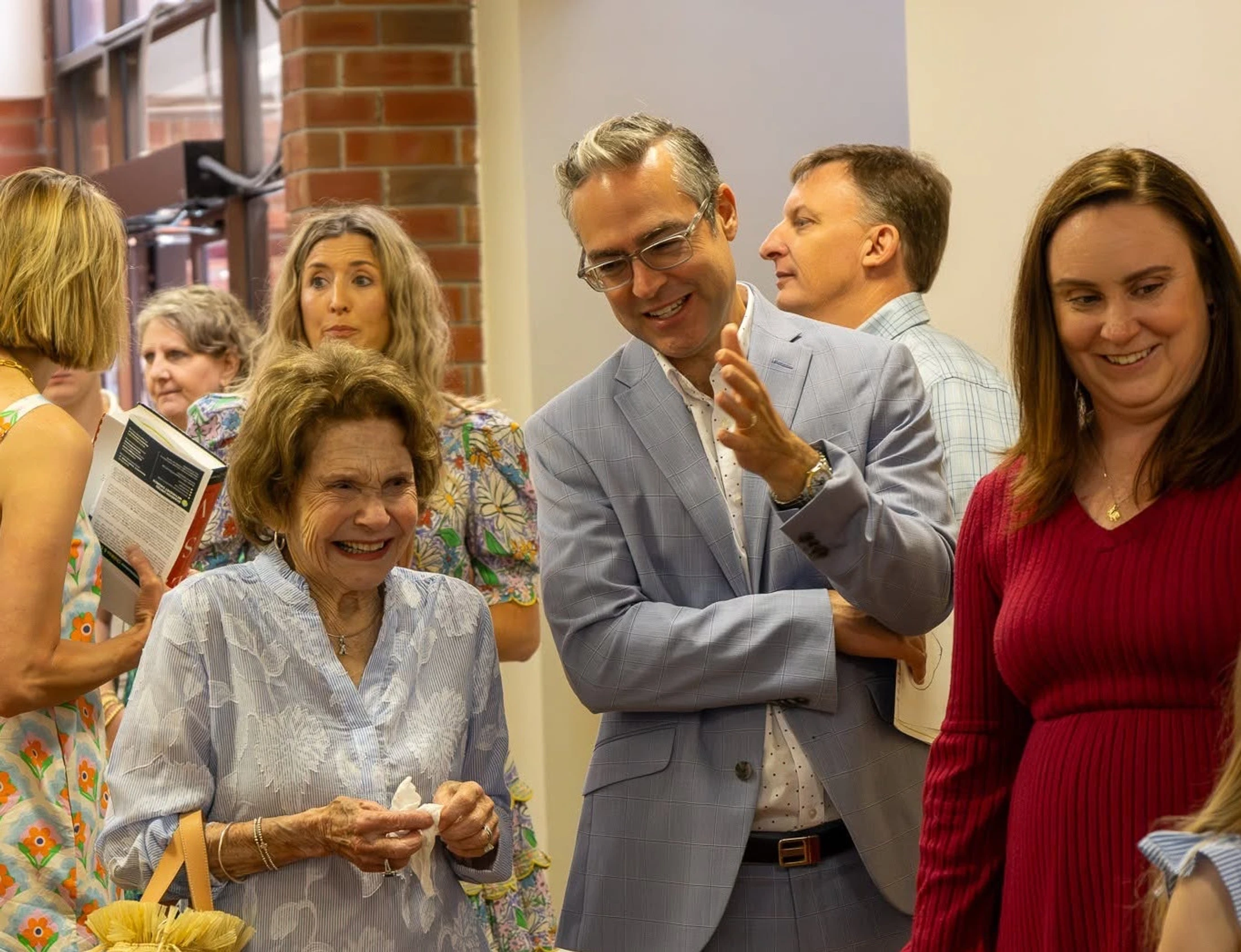 An older woman in a blue blouse smiles while talking with a man in a light blue suit. They are surrounded by other people at an indoor social event.