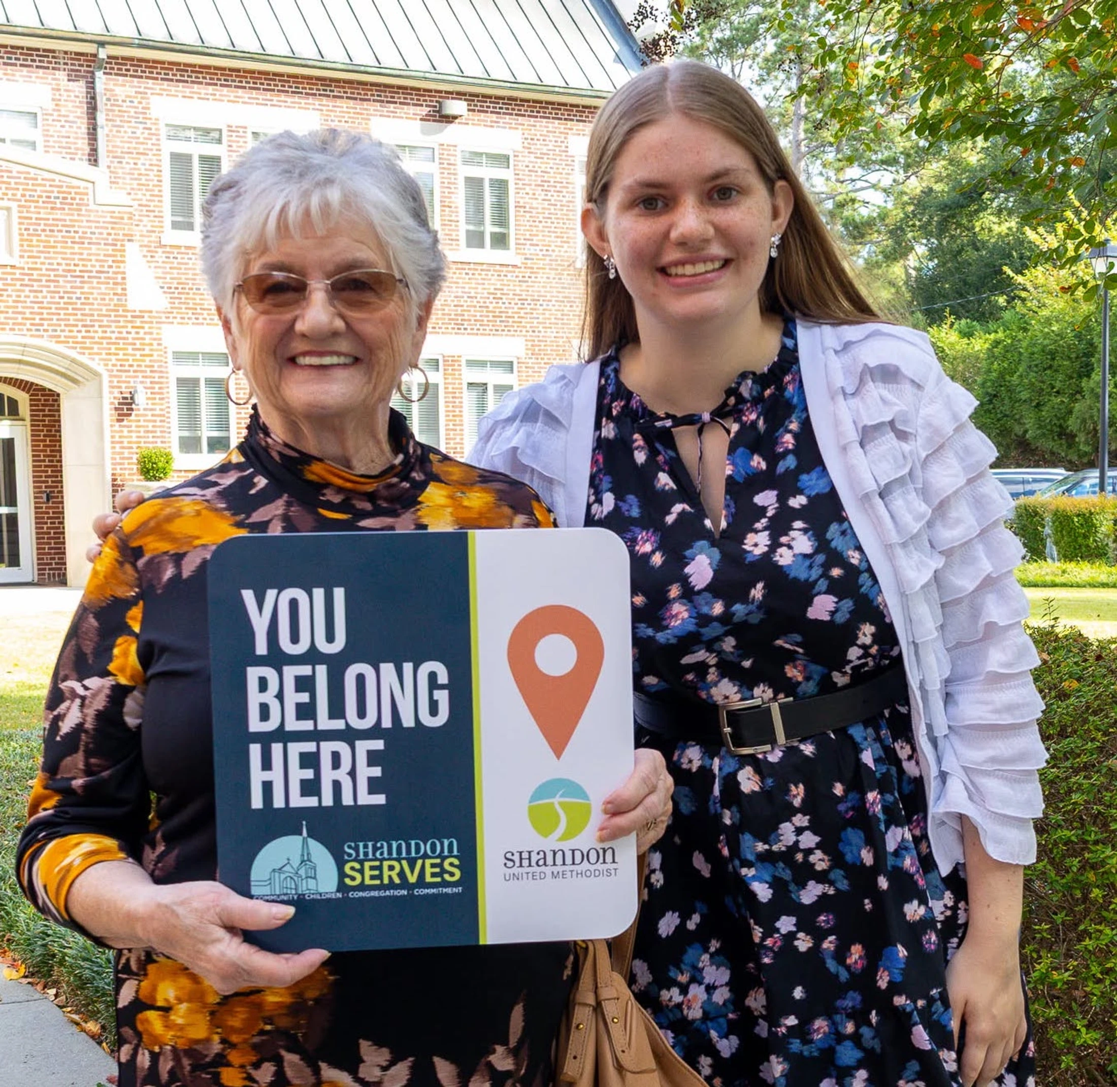 An older woman and a younger woman smile together outdoors. The older woman holds a sign that reads 'YOU BELONG HERE' for Shandon United Methodist church.