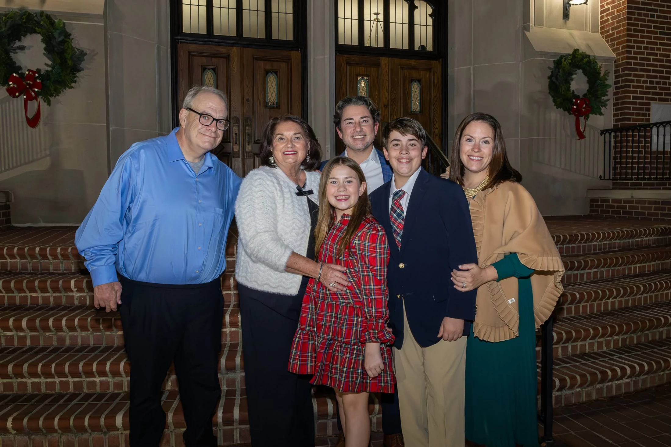 A smiling family of six, spanning three generations, poses for a photo on brick steps in front of a building decorated with Christmas wreaths at night.
