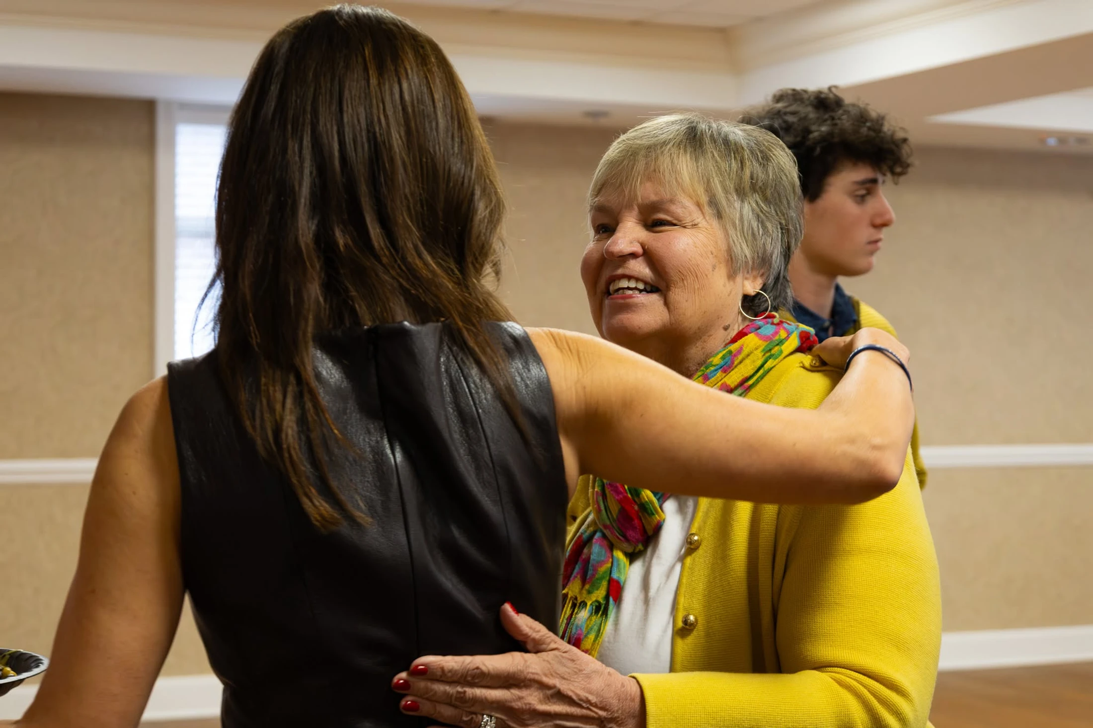 An older woman in a yellow cardigan smiles as she hugs another woman with brown hair. In the background, a young man with curly hair looks on.