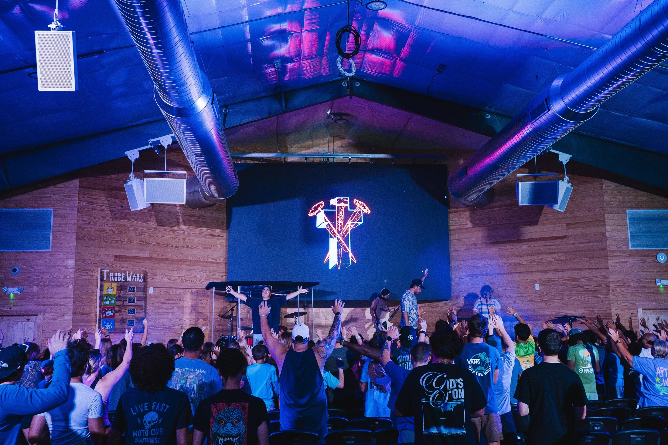 A large crowd of young people with their hands raised, watching a band on a stage under blue lights during a worship service in a large hall.