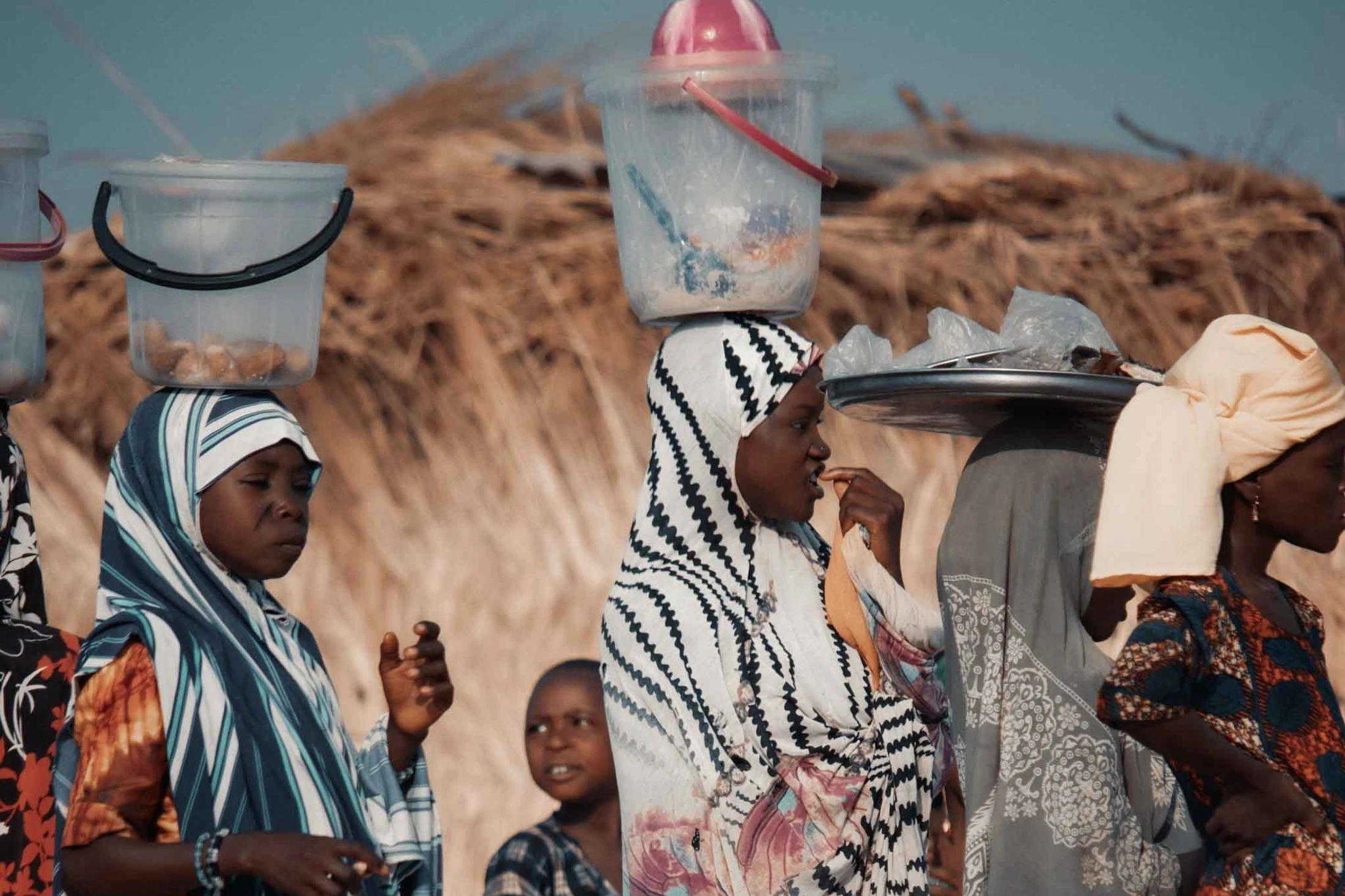 A group of African women in colorful headscarves carry buckets and trays on their heads. A young boy looks on in the background of a rural setting.
