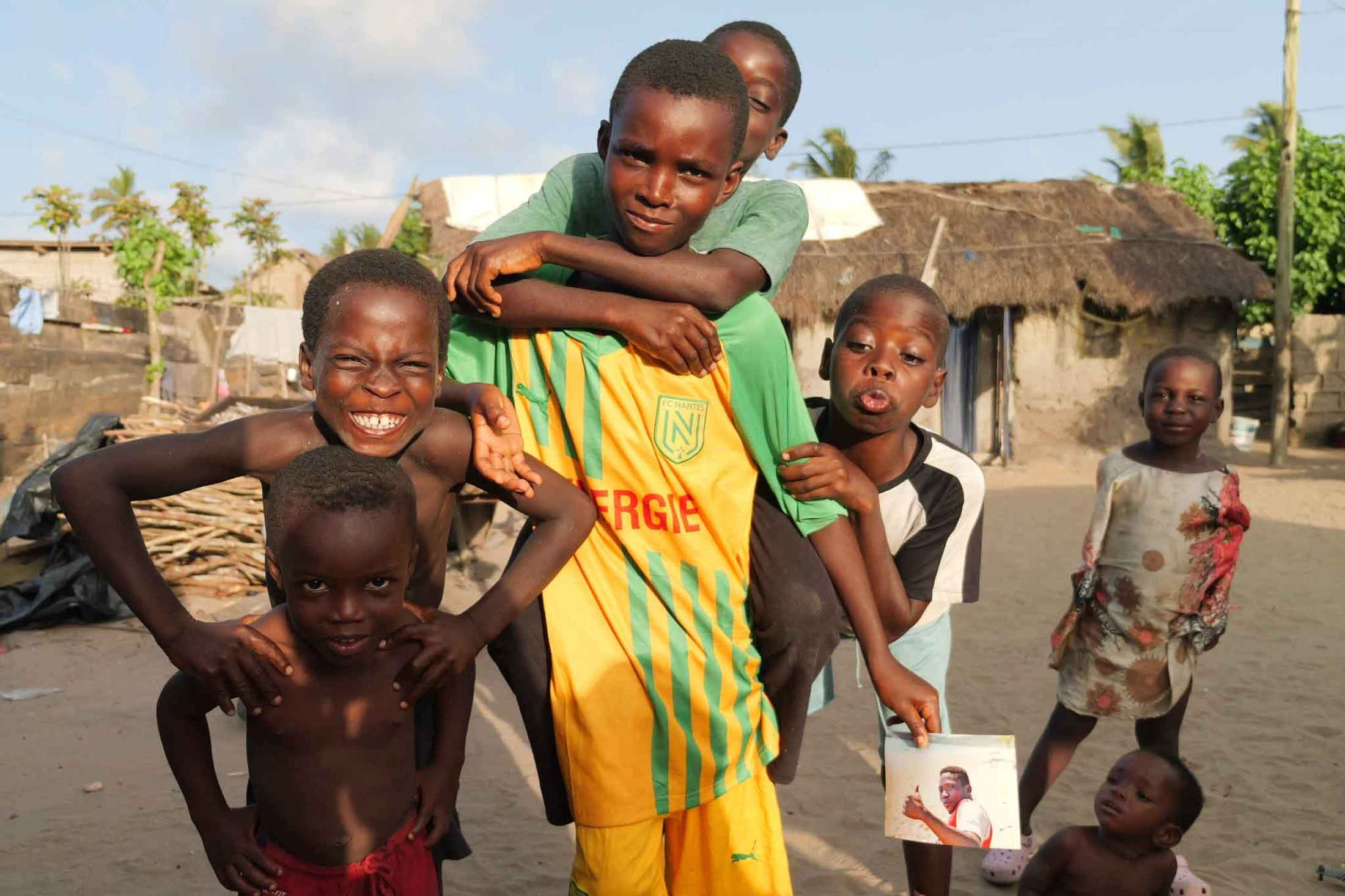 A group of young Black children happily pose together outdoors in a village. One boy carries another on his back, and they smile and make faces for the camera.