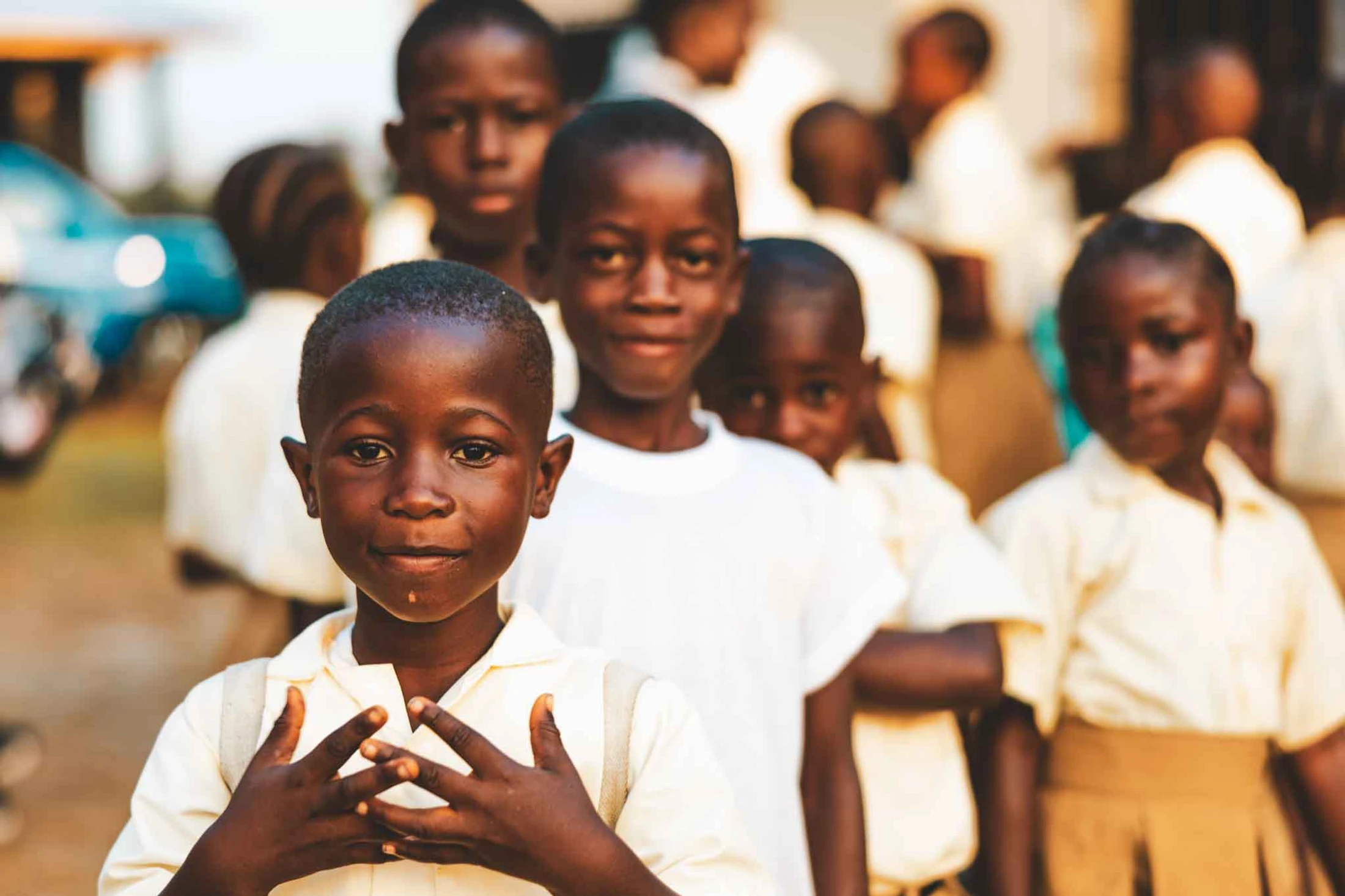 A young Black boy in a light-colored school uniform looks directly at the camera while standing in a line with other schoolchildren outdoors.