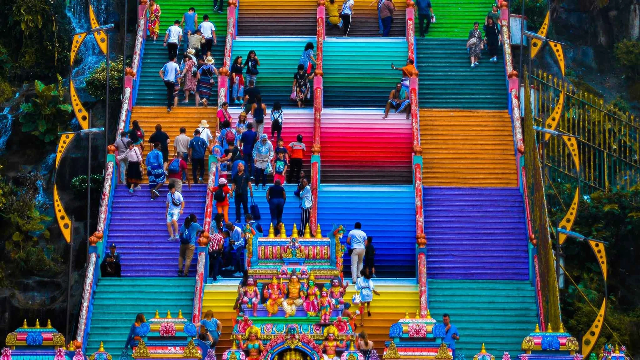 A high-angle view of many people climbing a massive, steep staircase painted in vibrant blocks of rainbow colors, leading up to a temple.