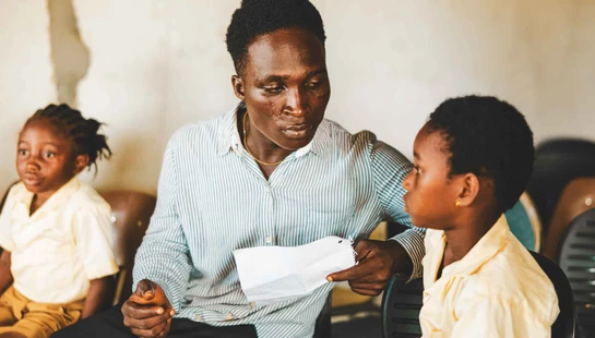 A Black man in a striped shirt sits and talks seriously with a young Black boy in a yellow shirt. Another young girl sits in the background.