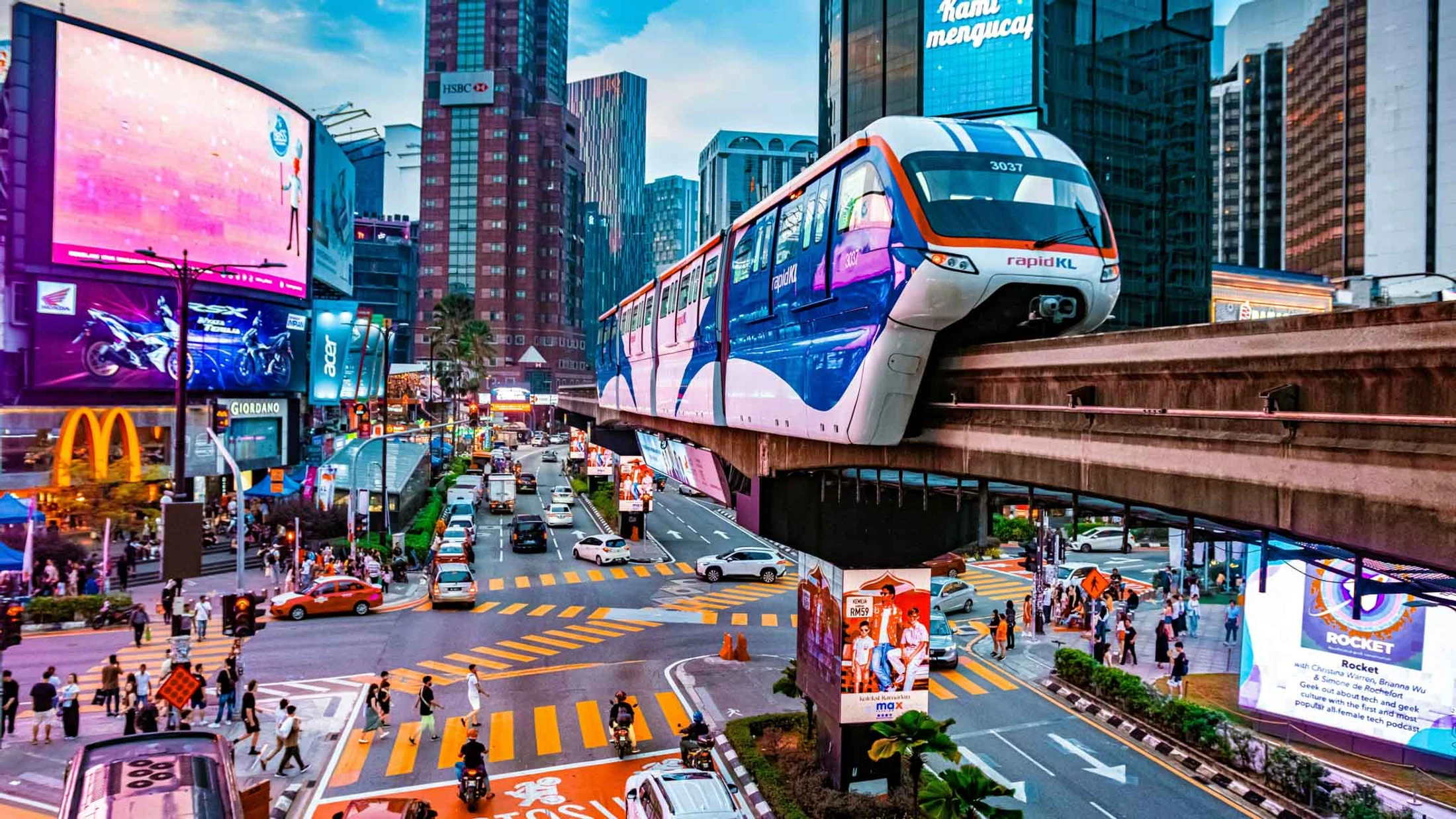 A modern monorail travels on an elevated track over a bustling city street in Kuala Lumpur, surrounded by tall buildings and vibrant digital billboards.