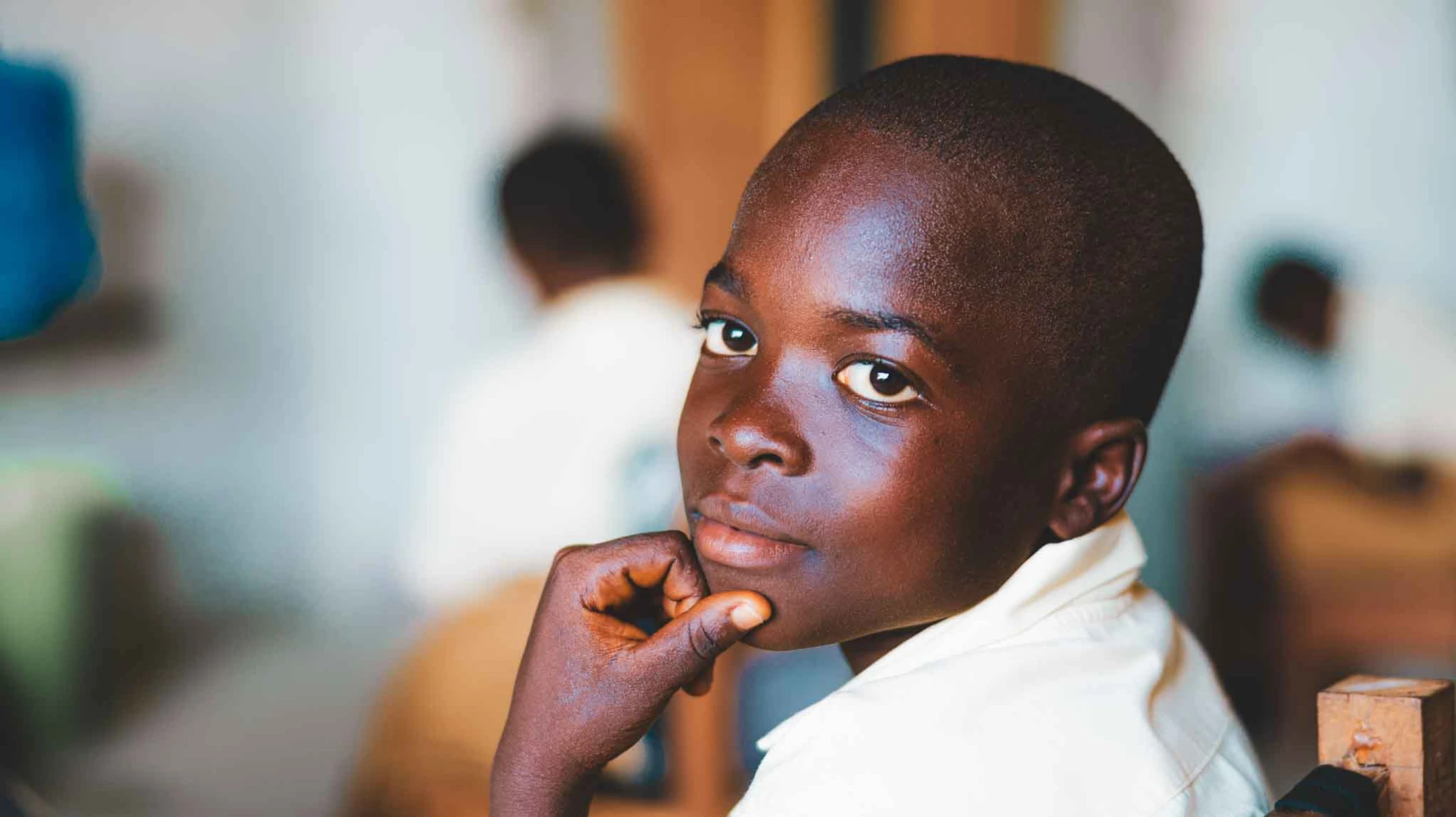 A young Black boy with short hair looks at the camera with a thoughtful expression, resting his chin on his hand. He appears to be in a classroom setting.